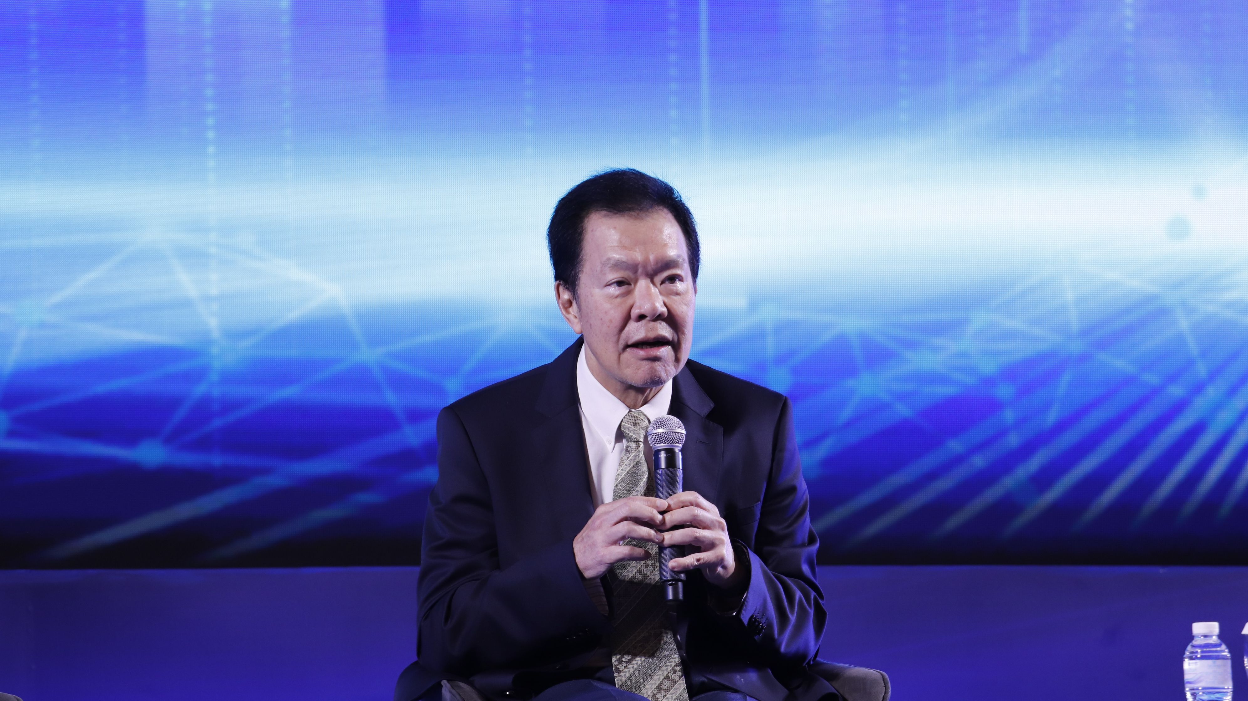 A man in a dark suit speaks into a microphone against a blue digital background.