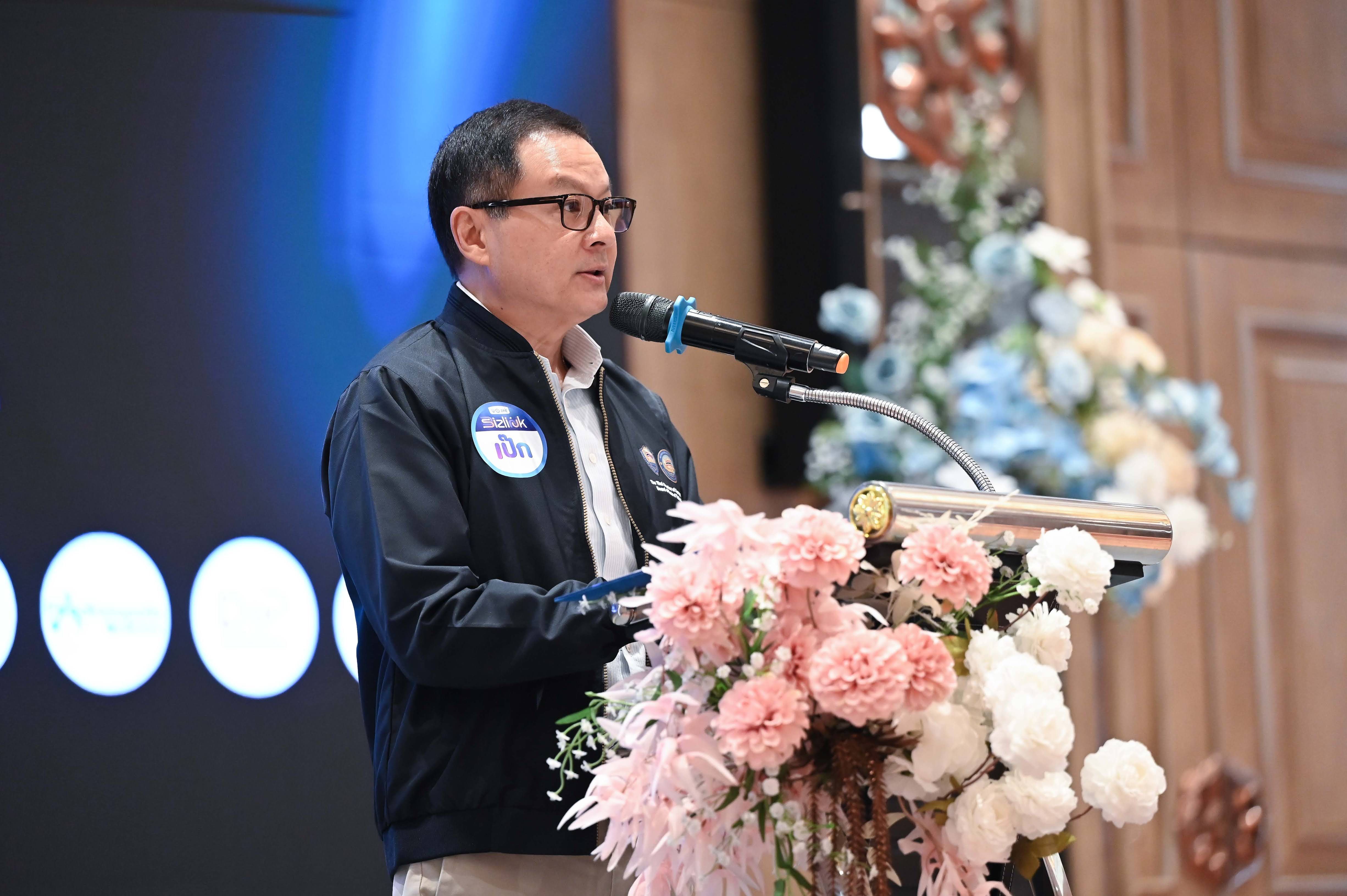 Man in glasses speaks at a podium with flowers, holding a microphone.