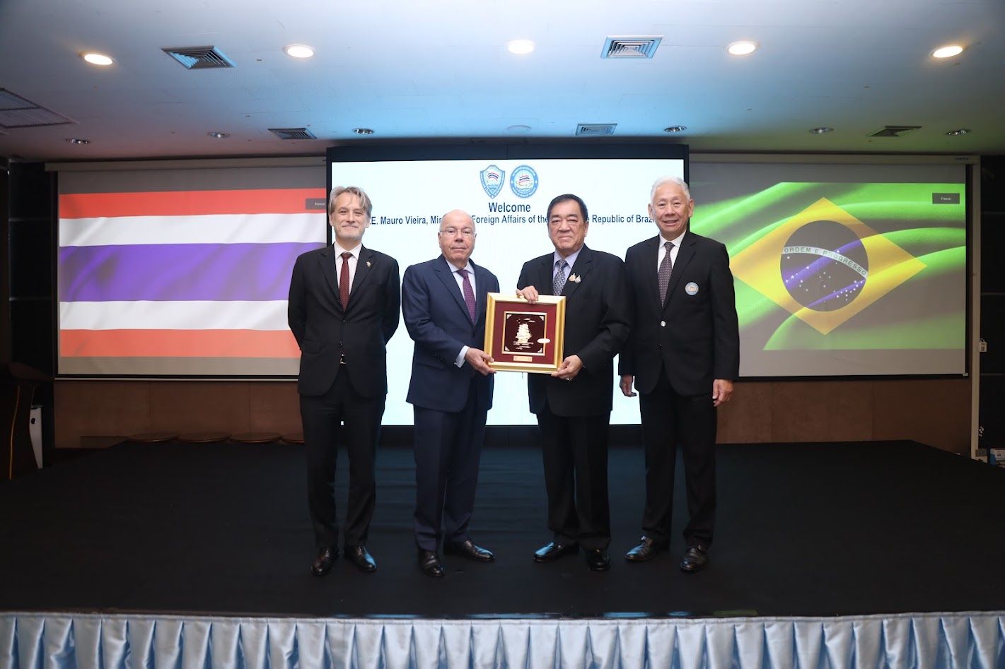 Four men in suits present an award on a stage with Thai and Brazilian flags.
