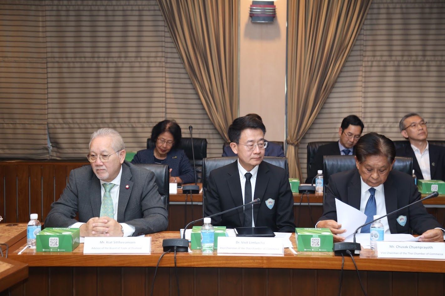 Three men in suits attend a formal meeting, one reviewing documents at a table.