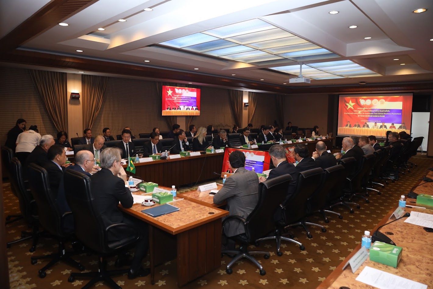 Attendees in a large conference room participate in a formal meeting with screens displaying presentations.