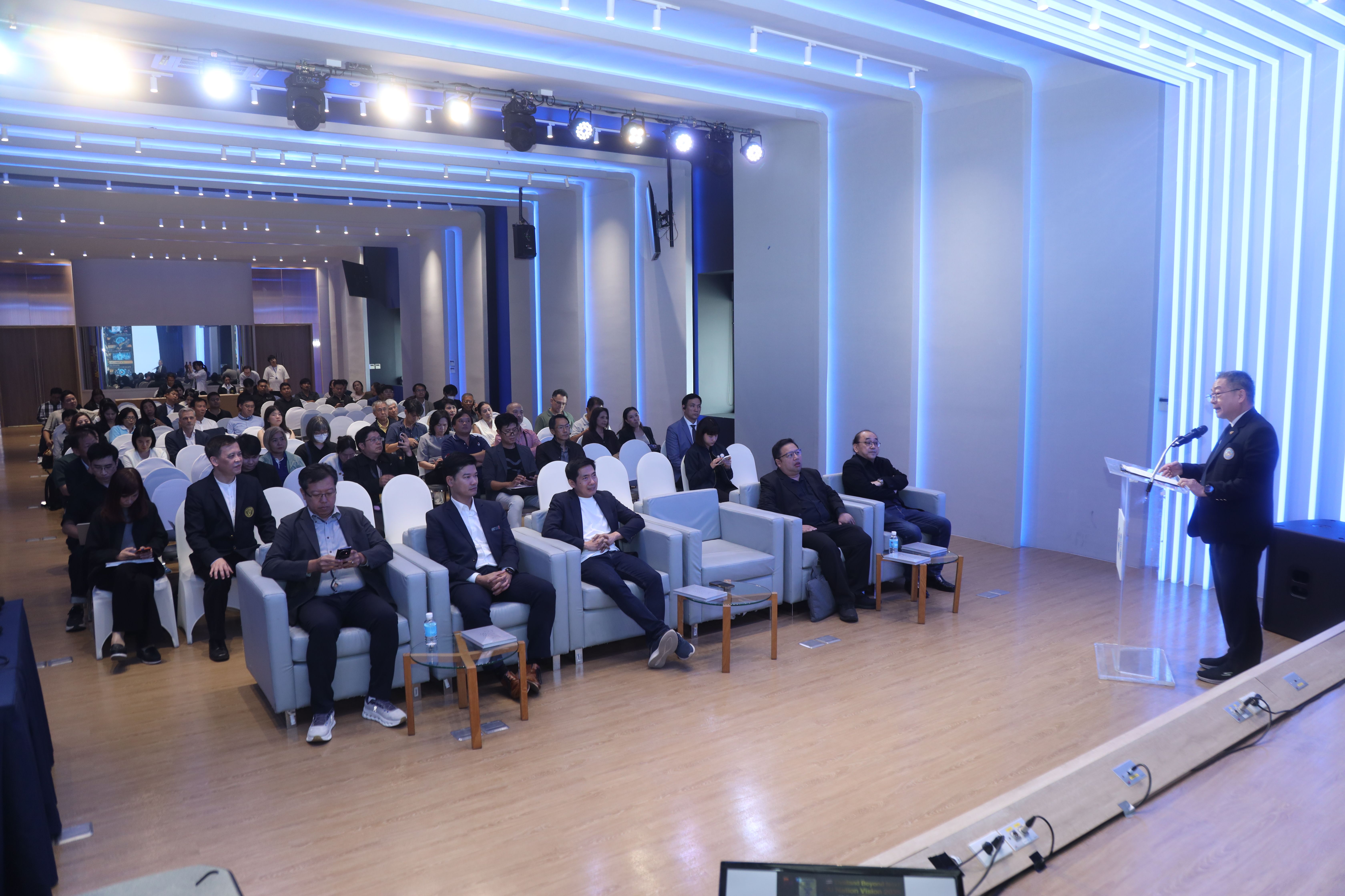A speaker addresses a large audience in a modern conference hall with blue ambient lighting.
