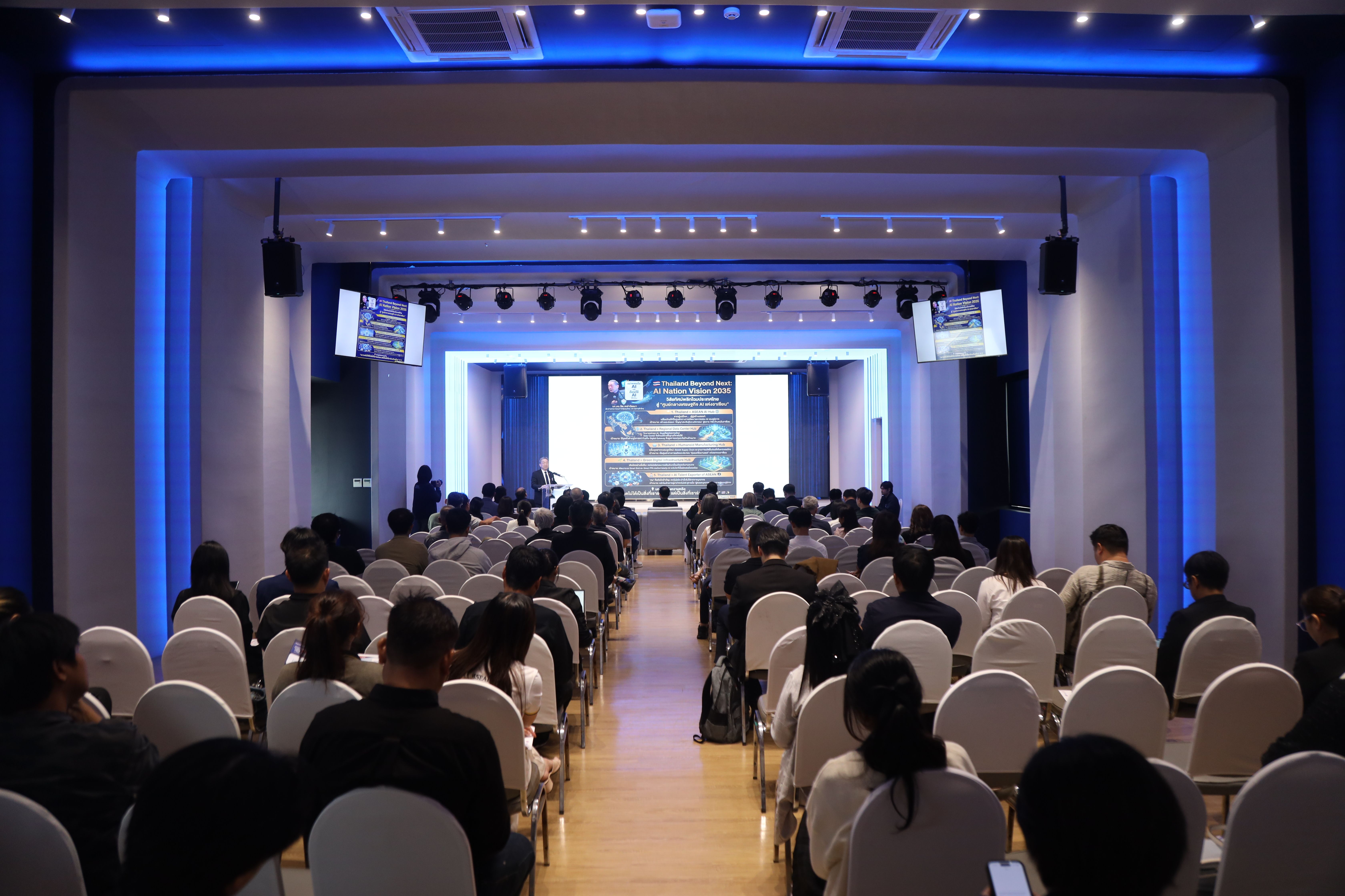 Audience in a modern conference hall, facing a stage with a large screen.