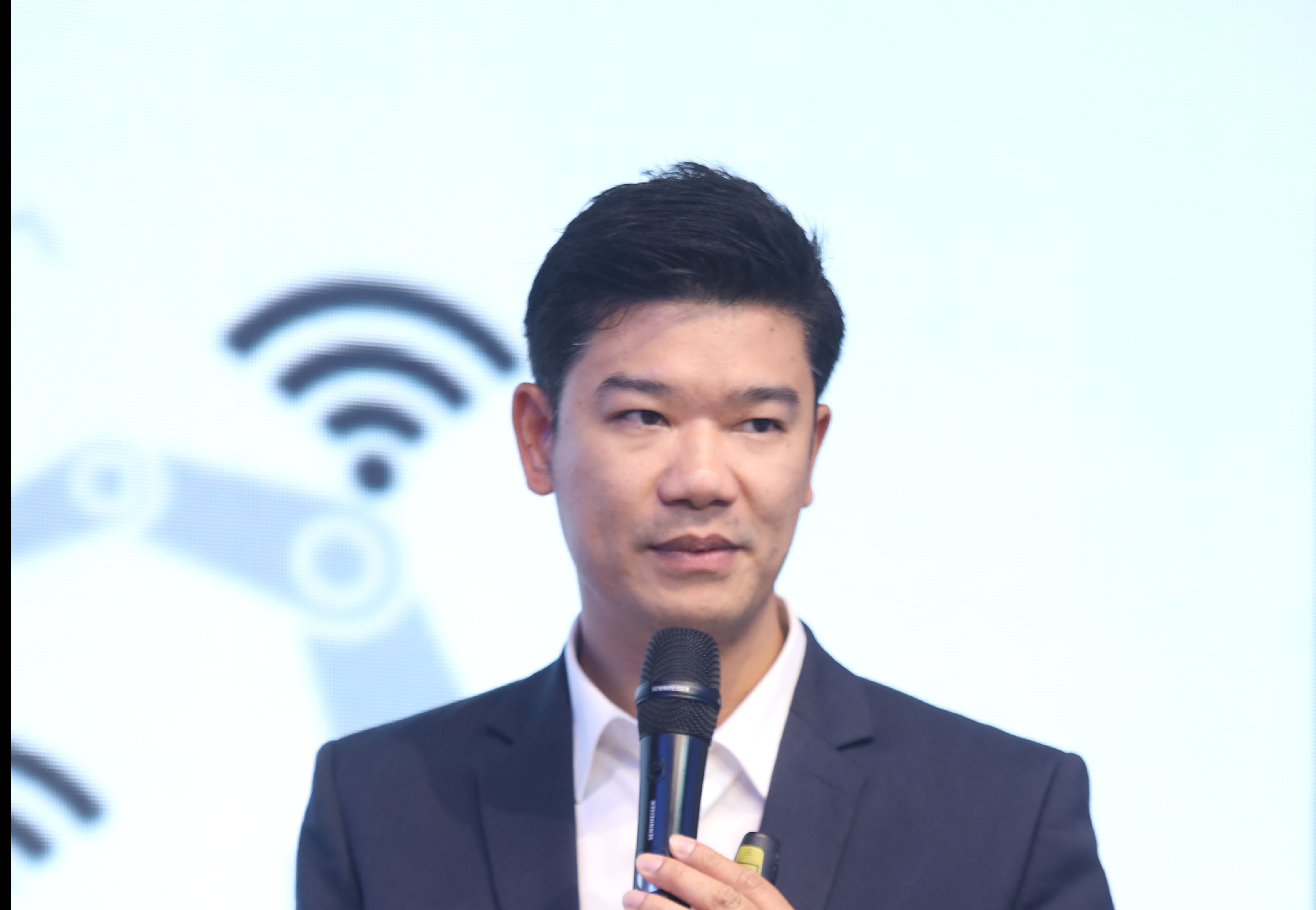 A man in a suit speaks into a microphone against a background of tech symbols.