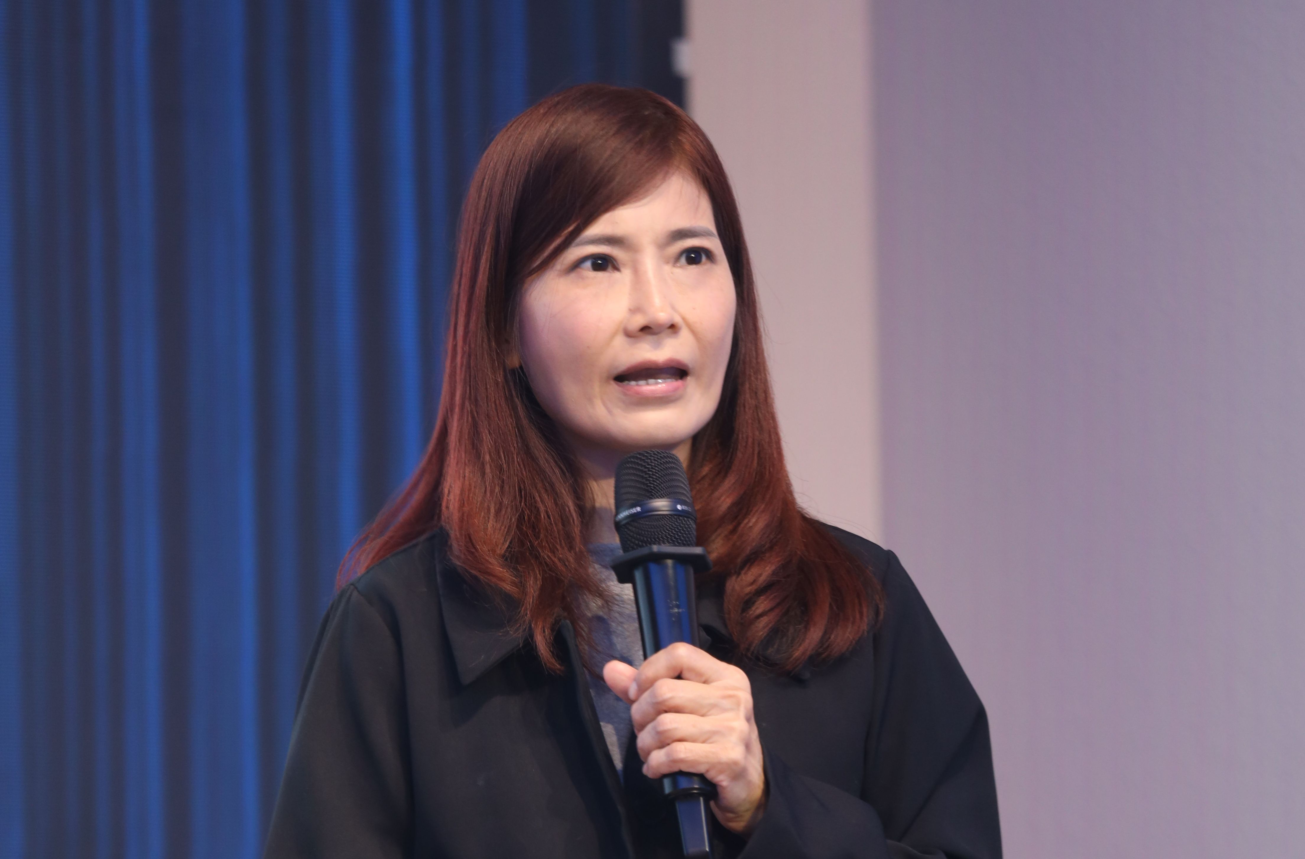 Asian woman with reddish-brown hair speaking into a microphone during a presentation.