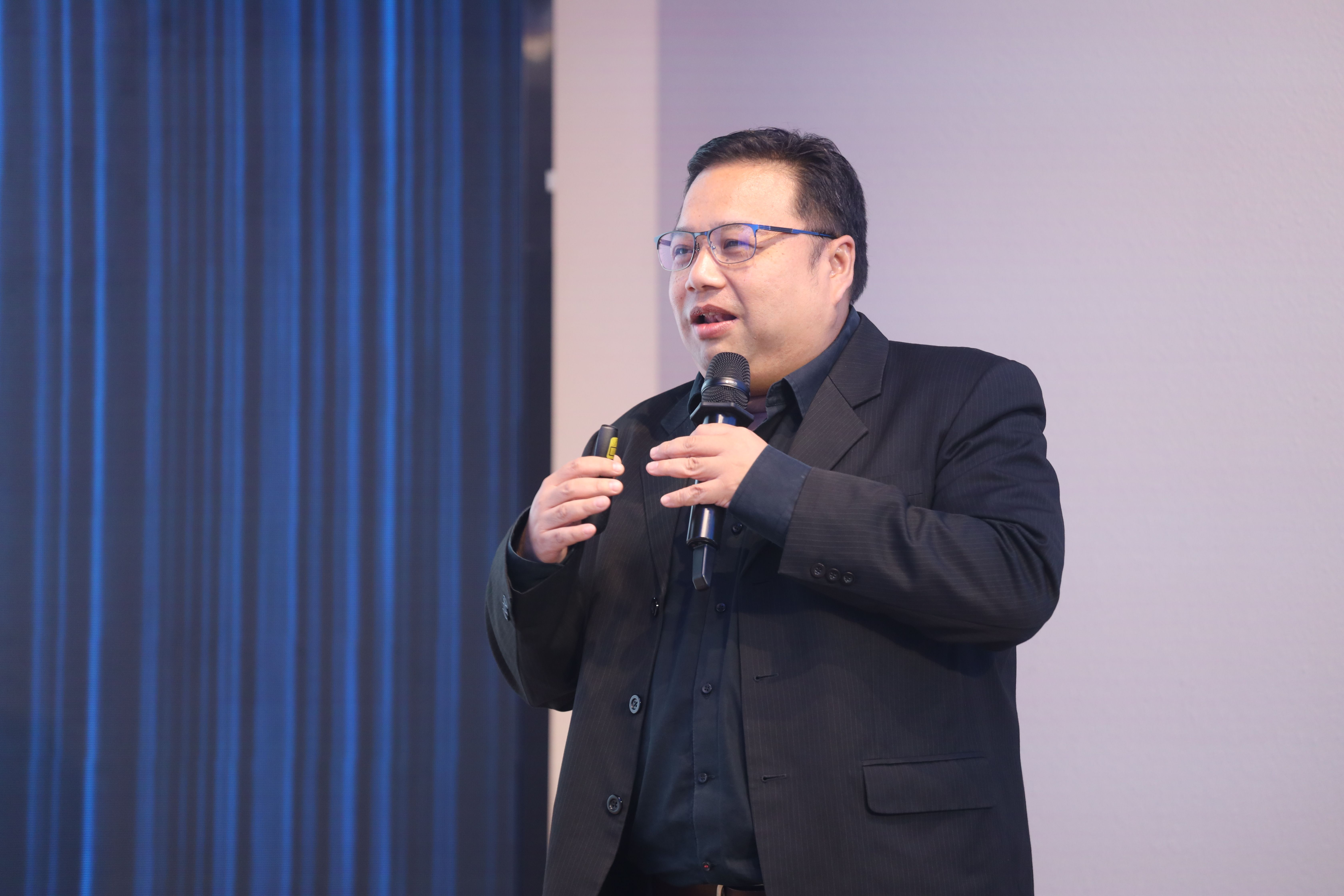 Man in a black suit and glasses speaks into a microphone during a presentation.