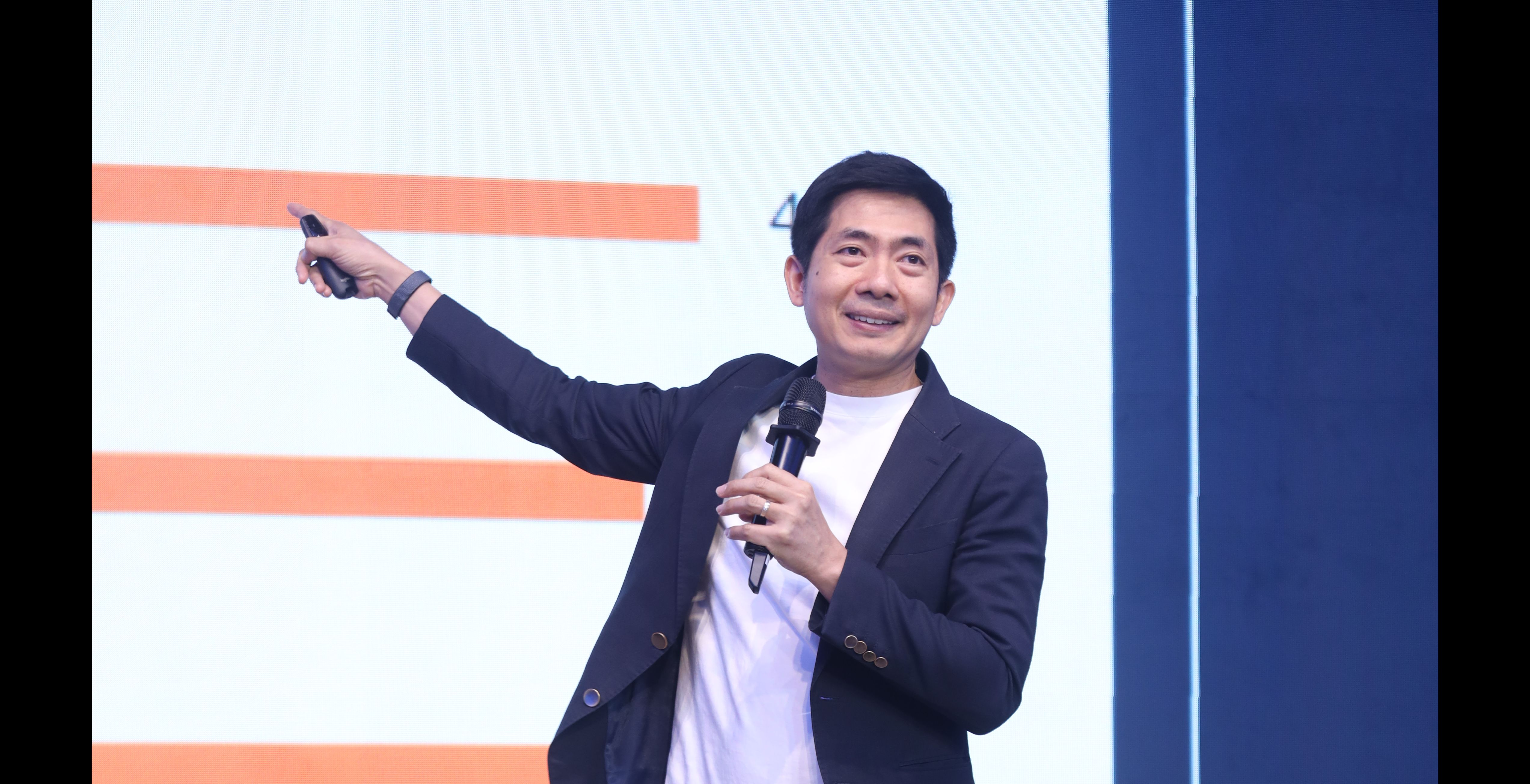 A smiling man presents on stage, holding a microphone and pointing with his other hand.