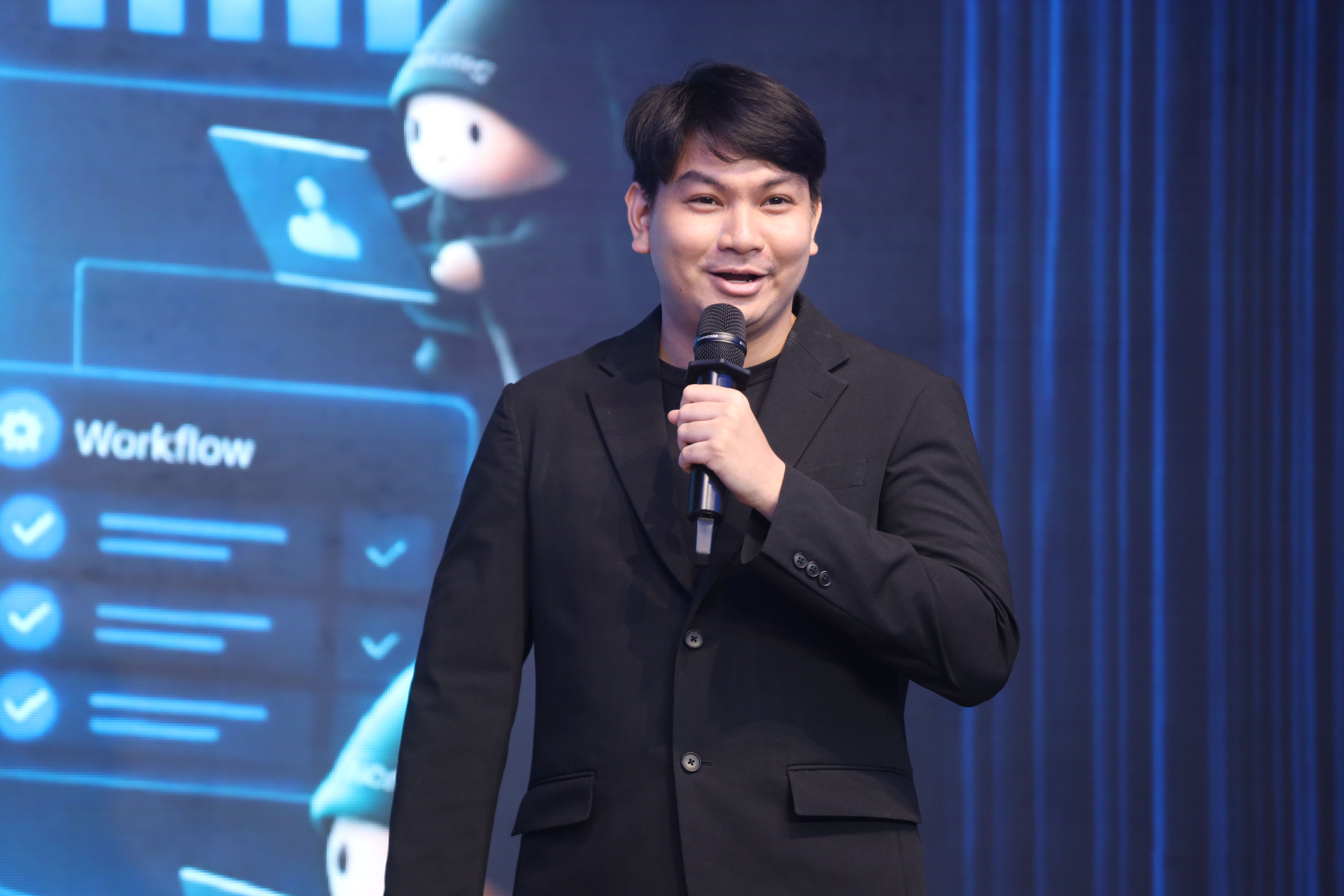 Man in black suit speaks into microphone before a screen showing workflow graphics.