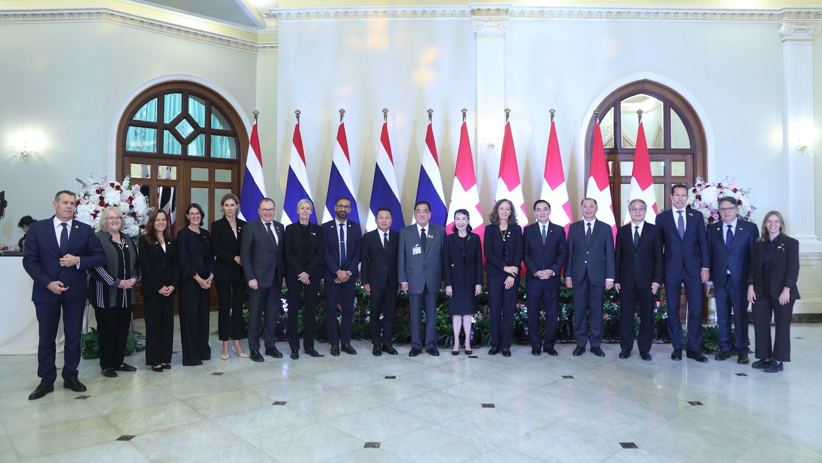 Group of officials stand before alternating Thai and Canadian flags in a formal room.