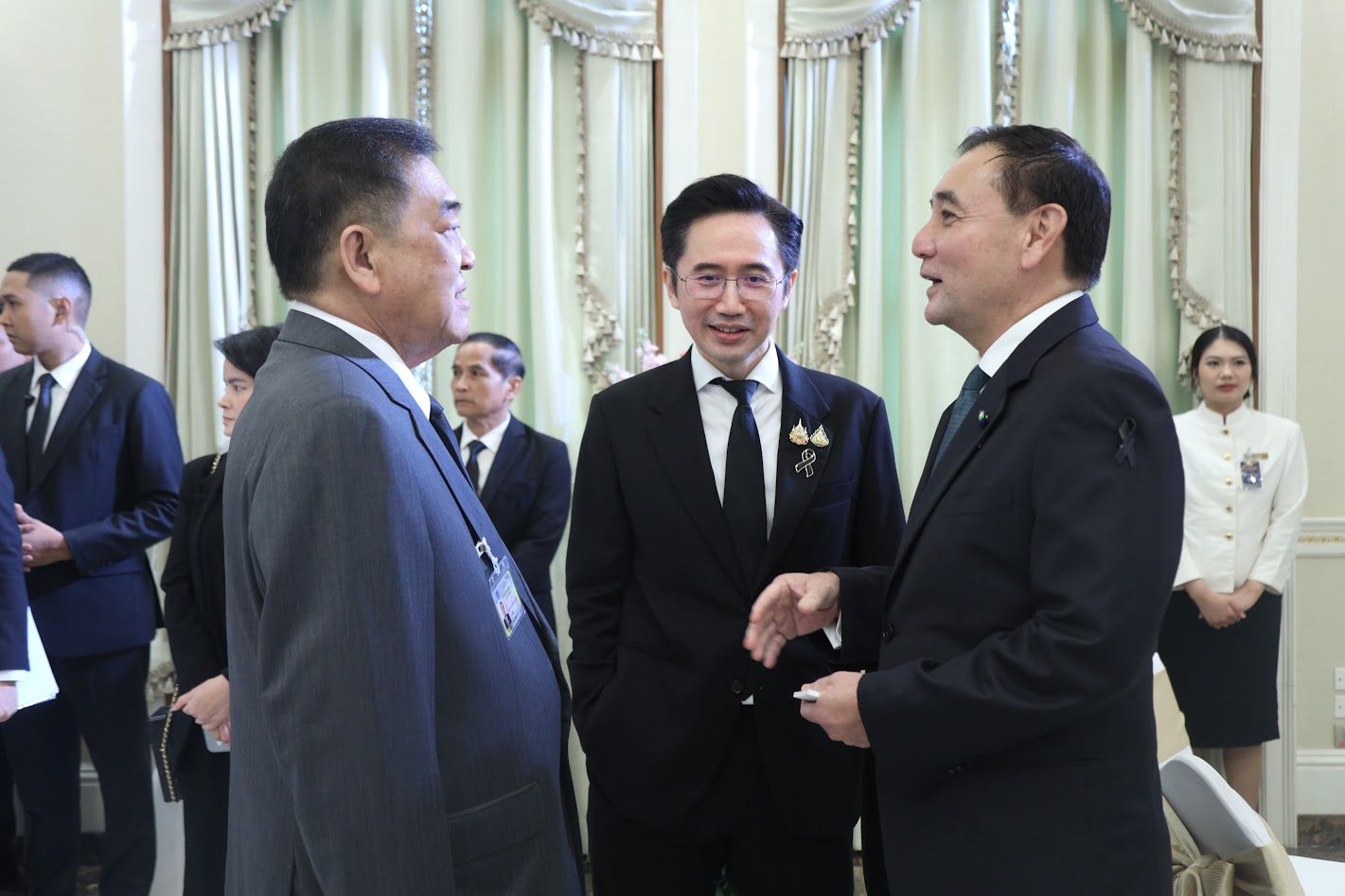 Three men in dark suits conversing formally in an elegant indoor setting.