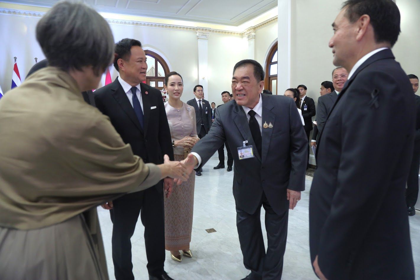 An older woman shakes hands with a man in a dark suit at a formal indoor event.