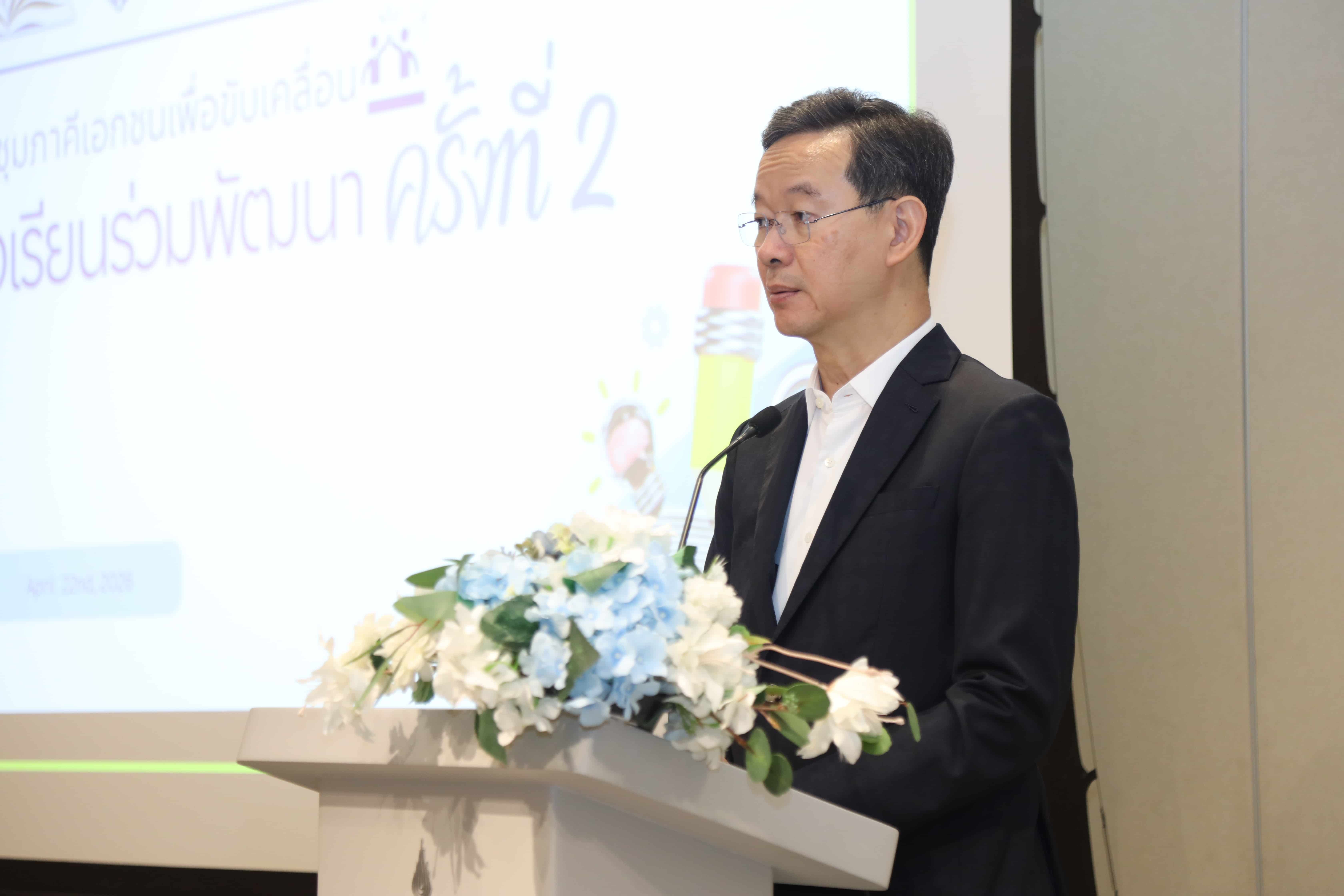 Man in a suit speaks at a podium with flowers and a screen showing Thai text.