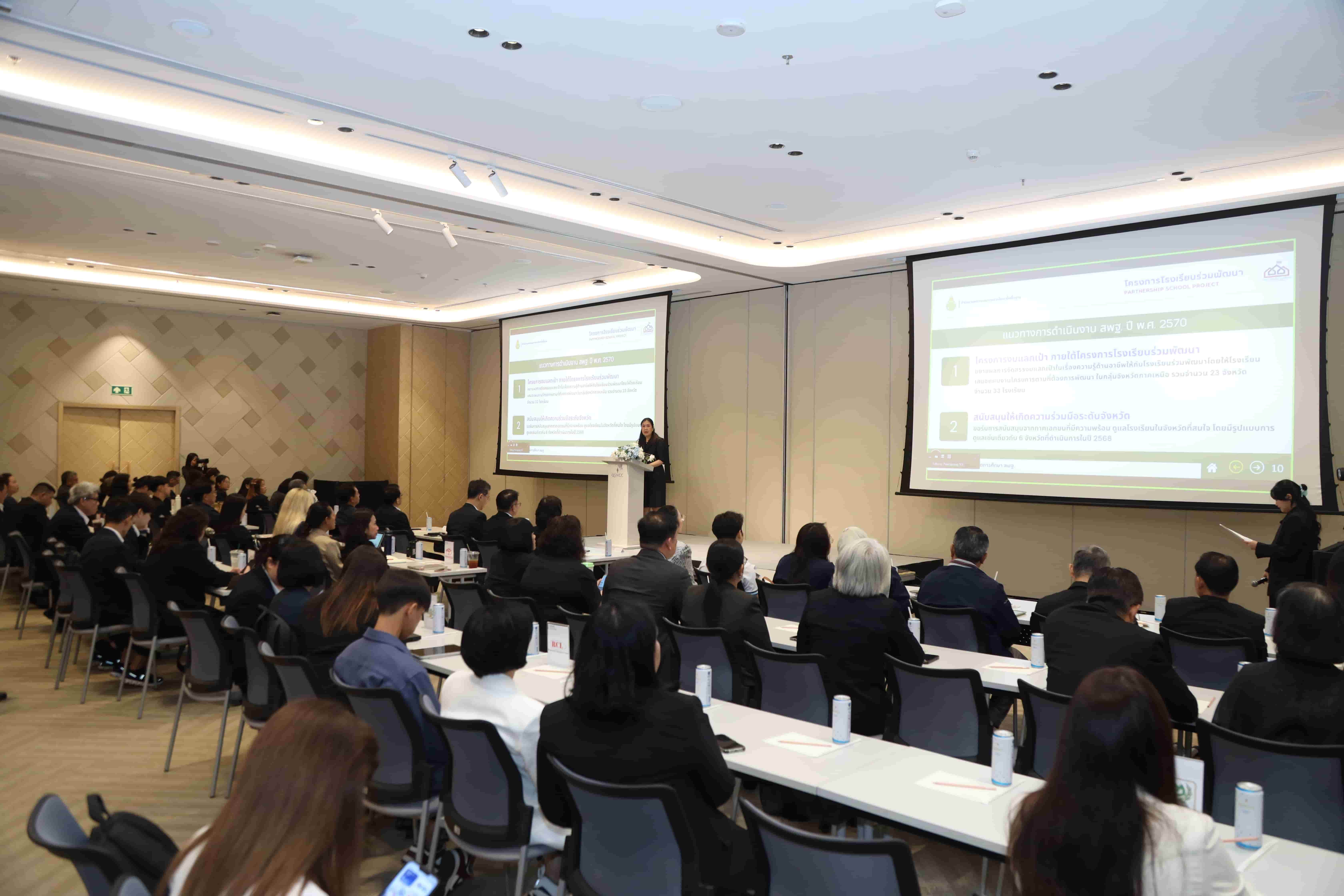 Attendees observe presentations on large screens in a brightly lit conference room.