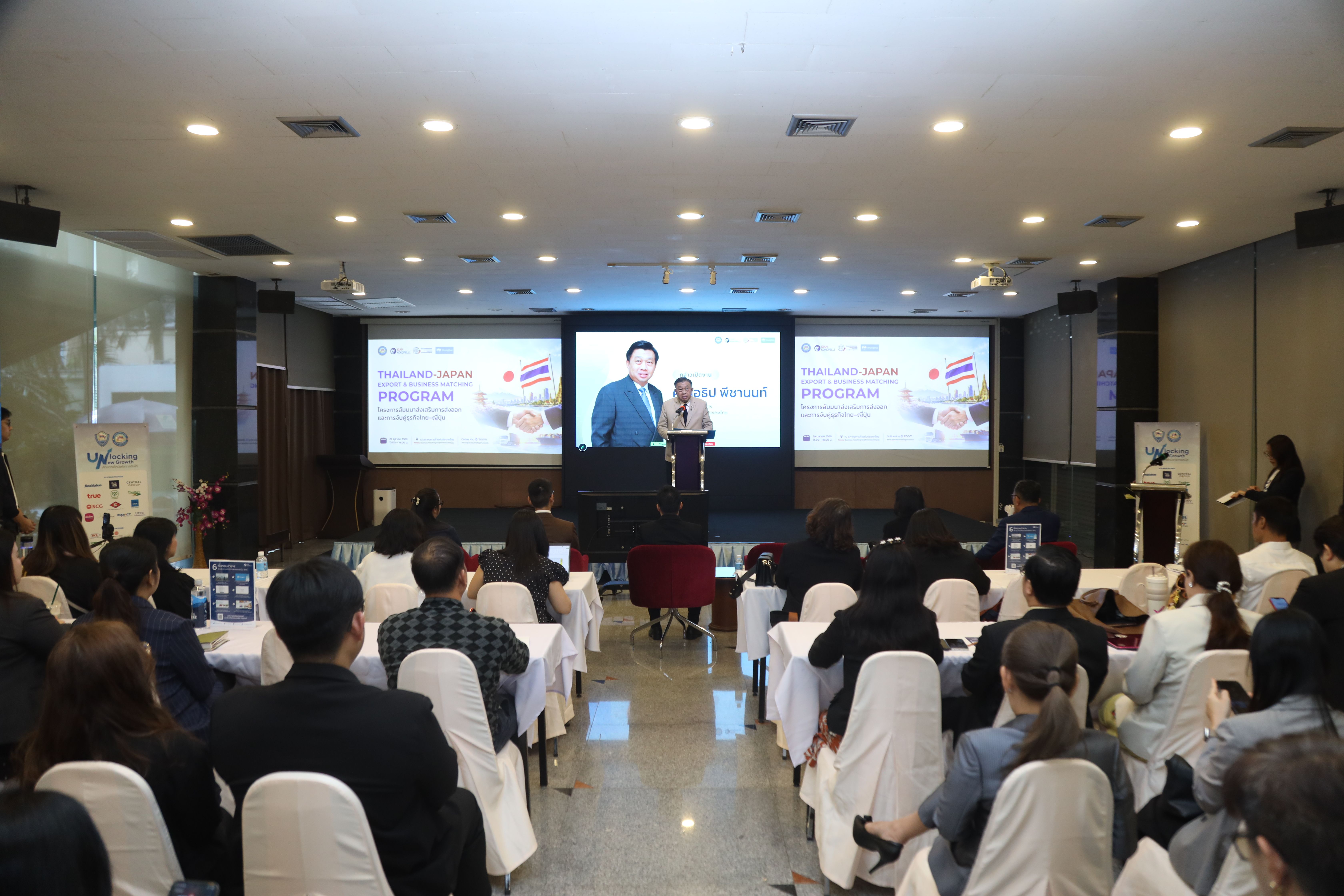 A speaker presents on a brightly lit stage to an audience in a formal conference room.