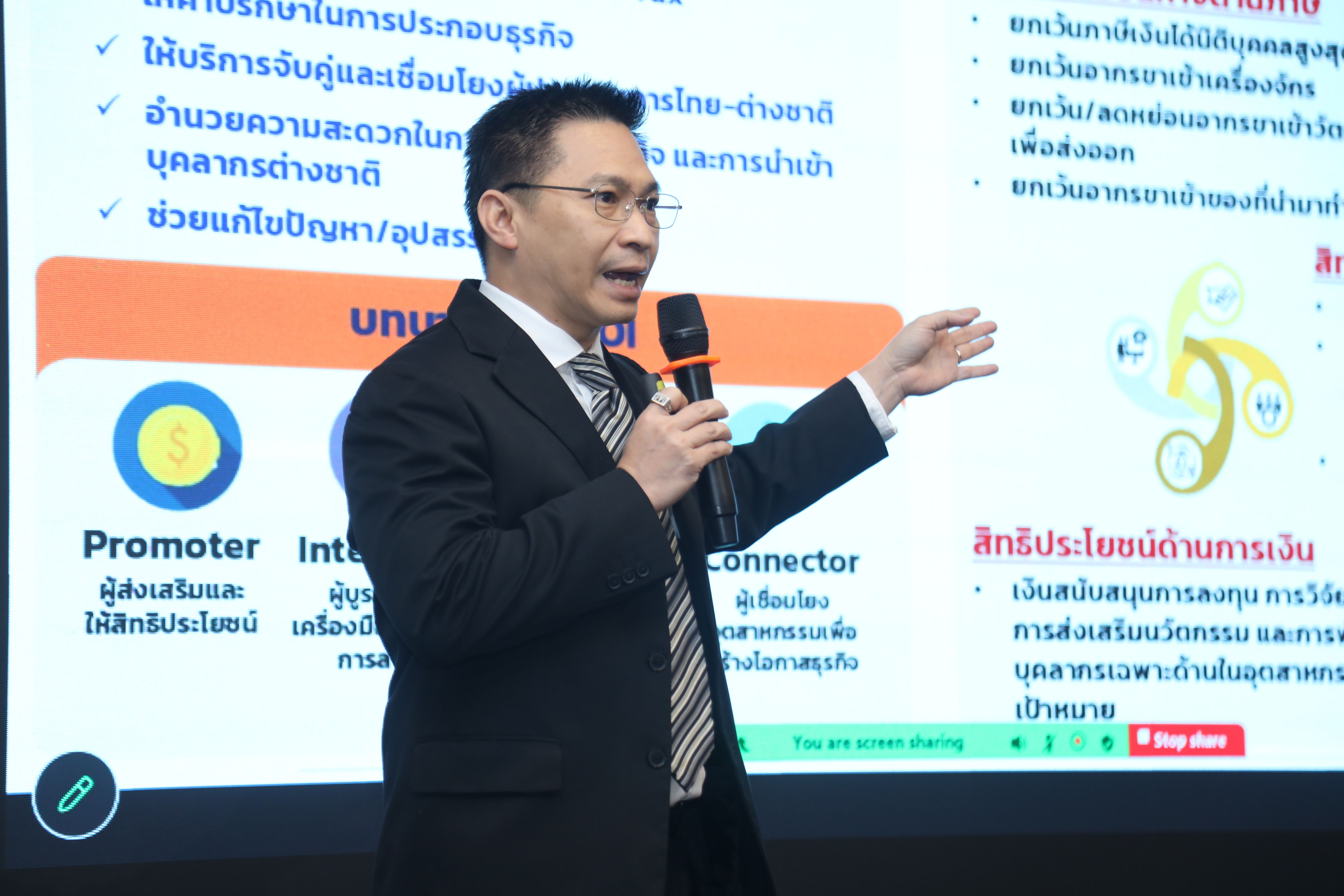 A man in a suit and glasses presents, gesturing to a screen with text.