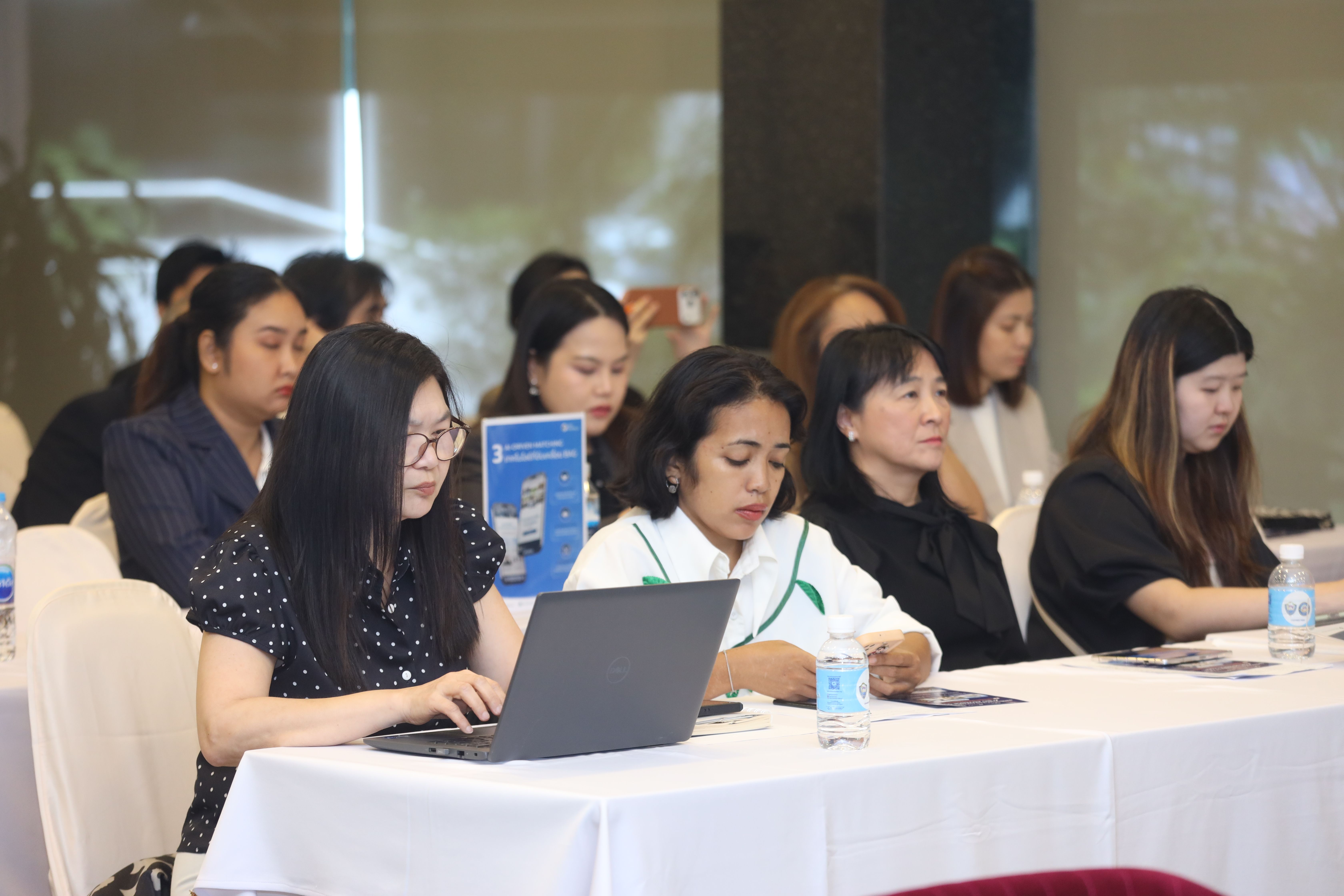 Professional women at a conference, one actively typing on a laptop while others listen.