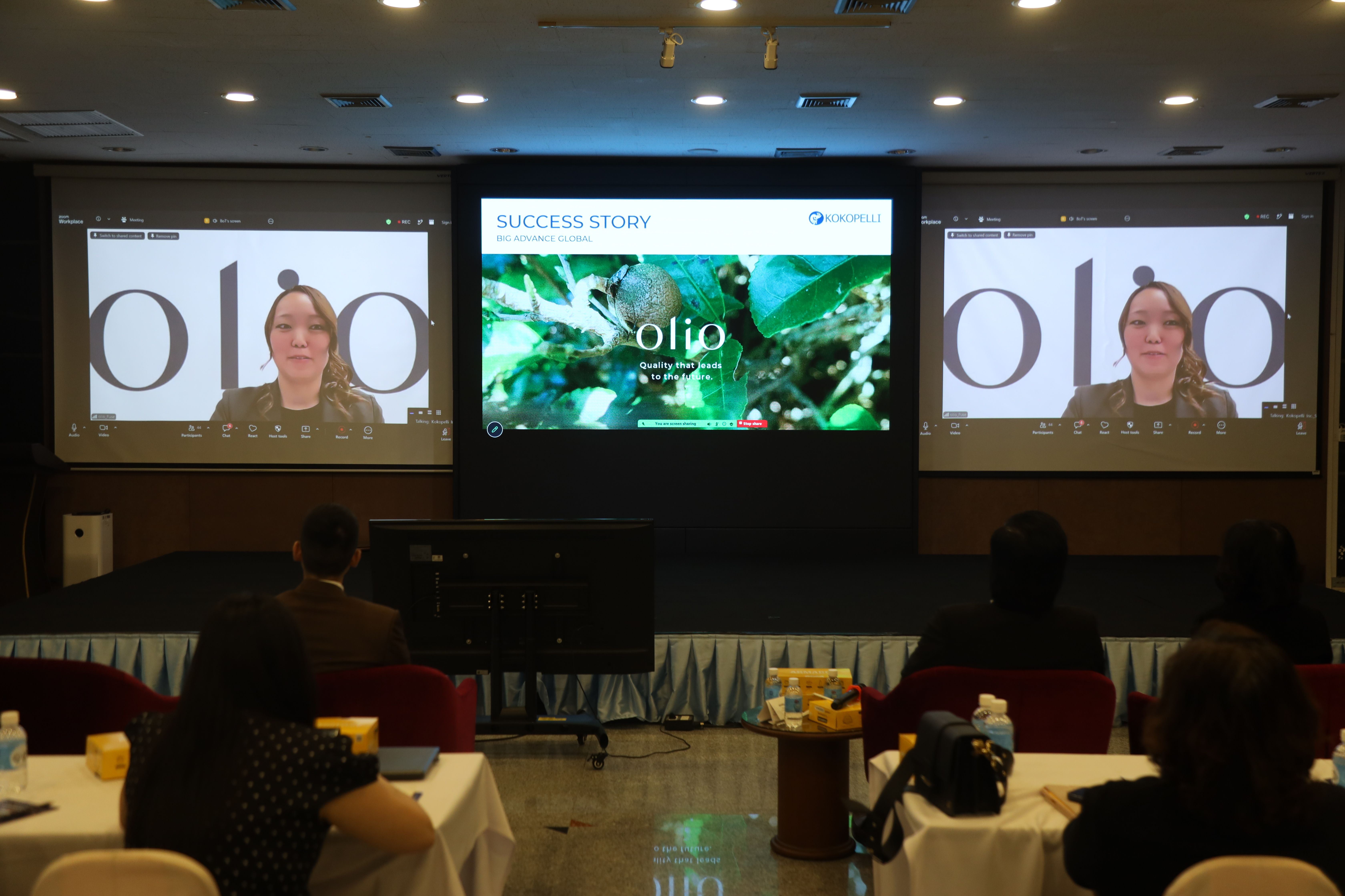 A conference audience views large screens, one showing 'Solio' and others a video call.