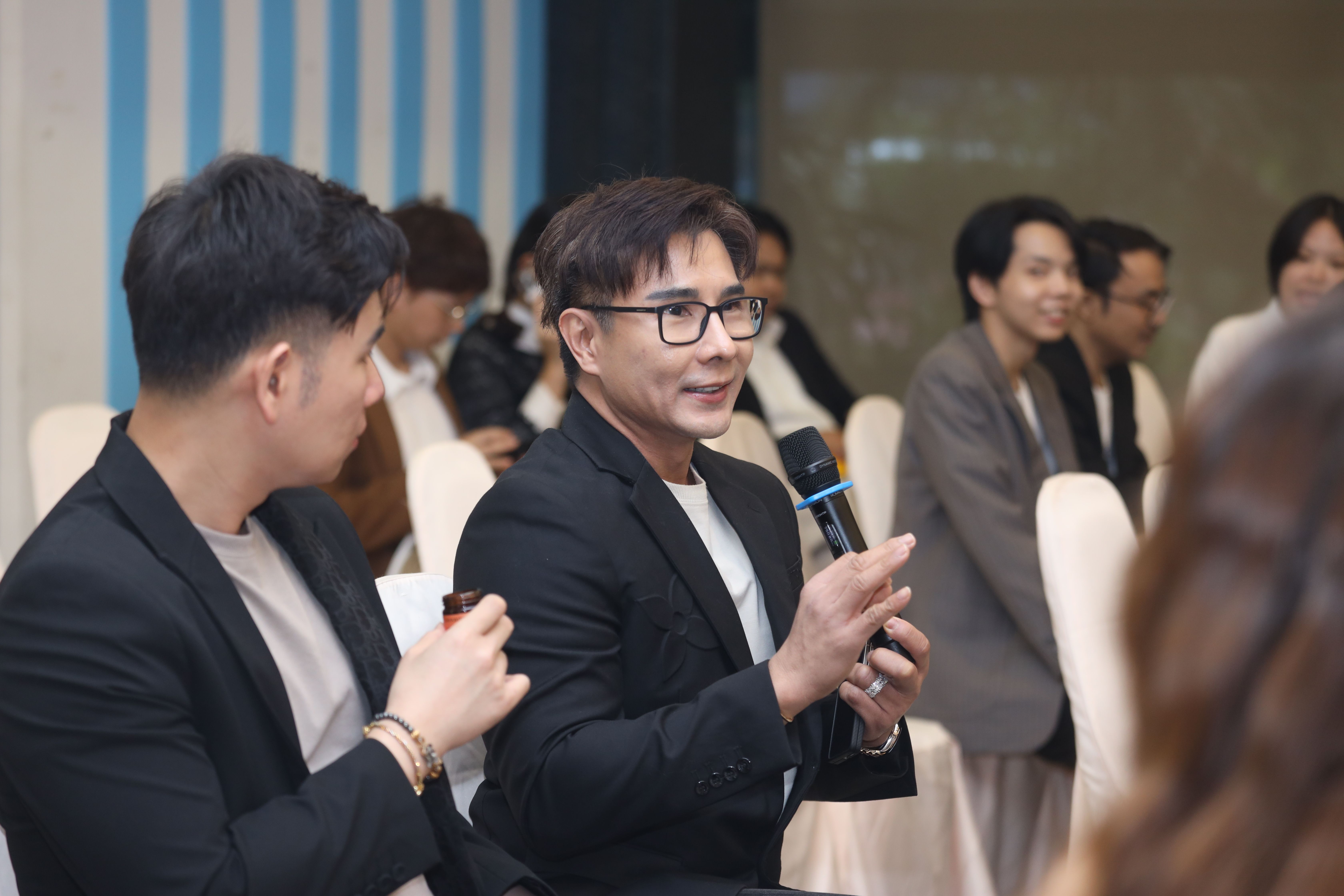 Man in glasses speaking into a microphone at an indoor event with other attendees.
