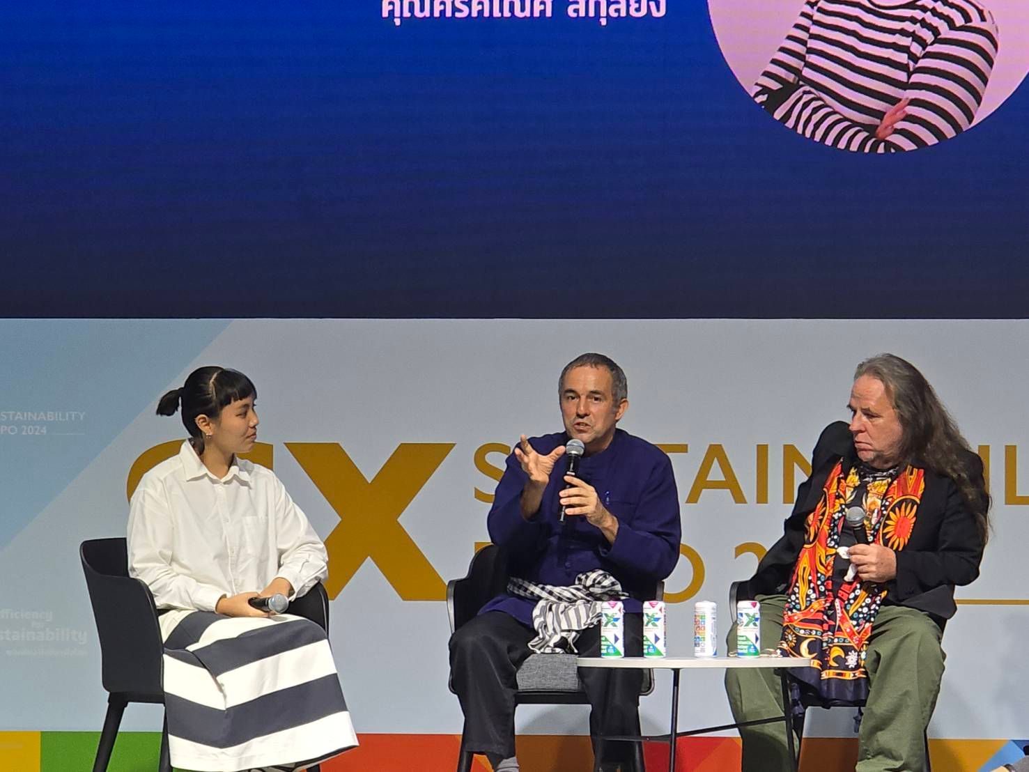 Three panelists discuss sustainability on a stage with a large screen behind them.