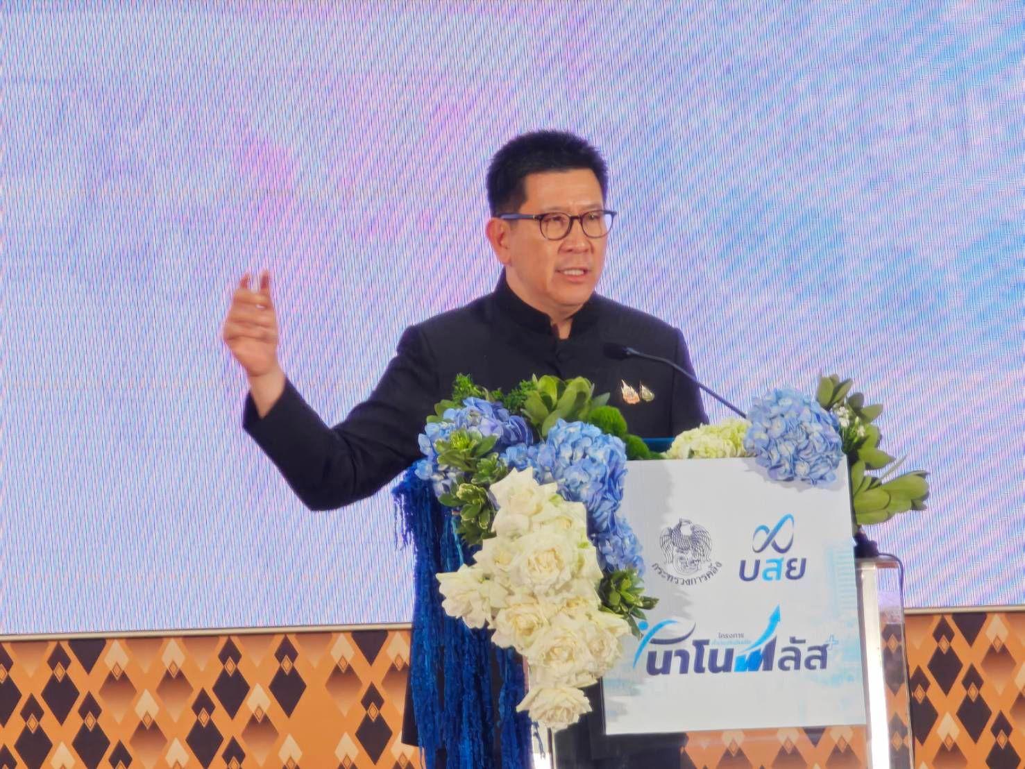 Man in dark suit and glasses speaking at a podium decorated with flowers and UOB logo.
