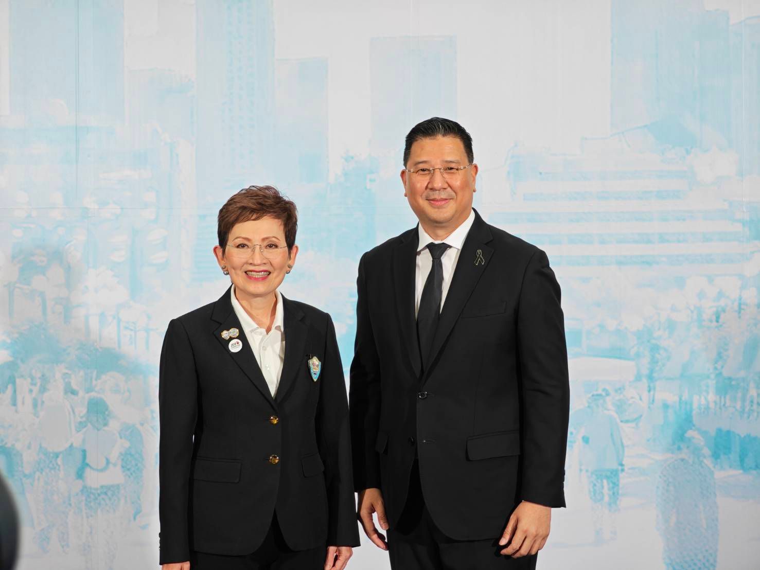 Woman and man in dark suits stand side-by-side, smiling against a light background.