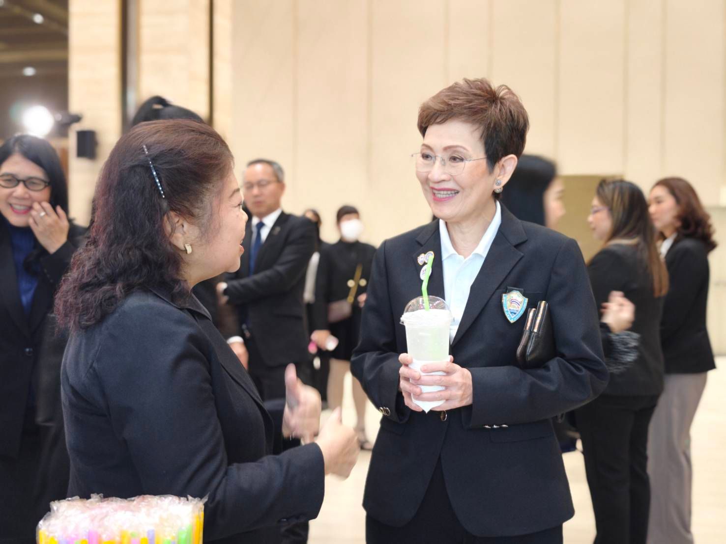 Two women converse in dark suits, one smiling and holding a drink.