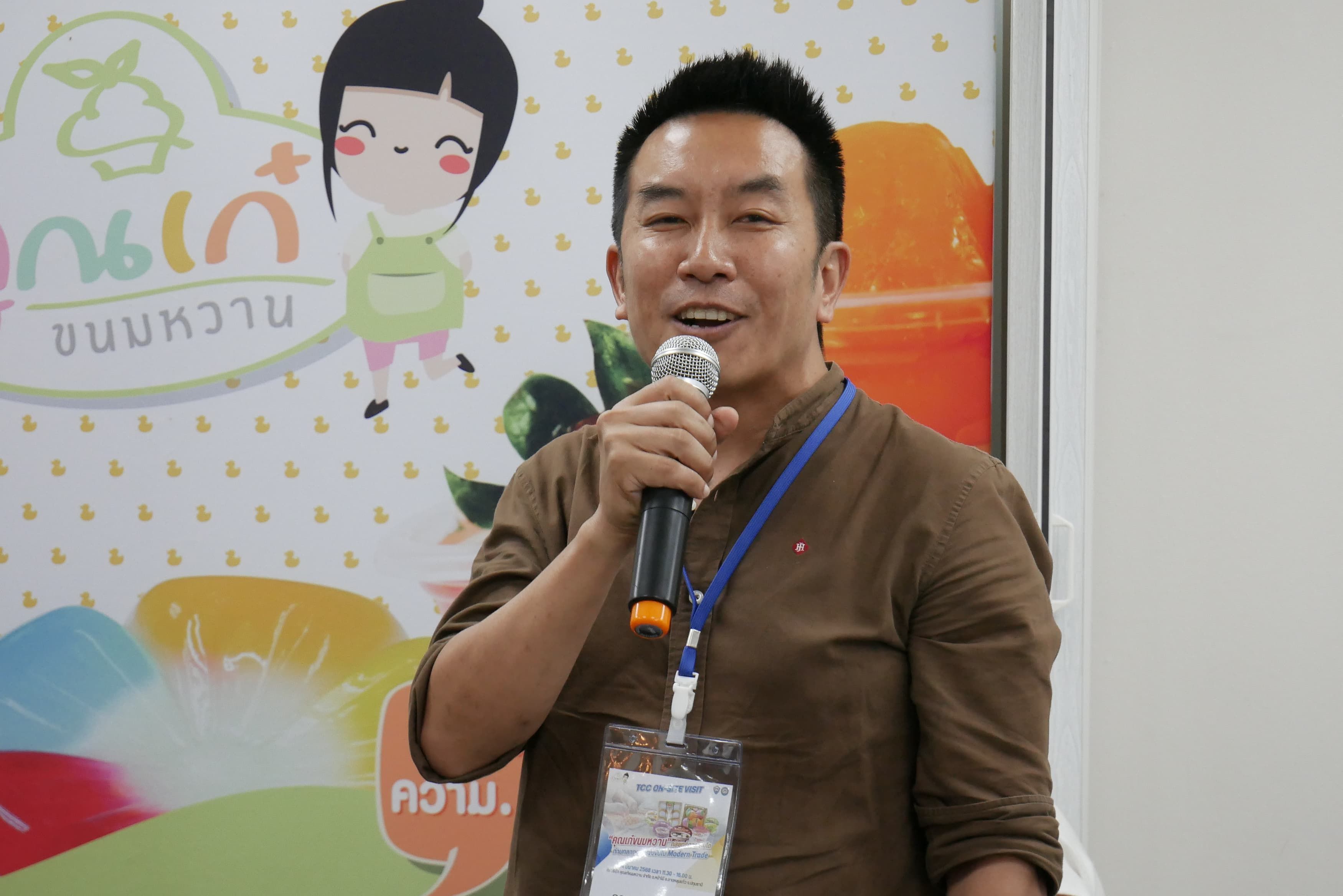 Man in brown shirt speaking into a microphone with a colorful cartoon background.