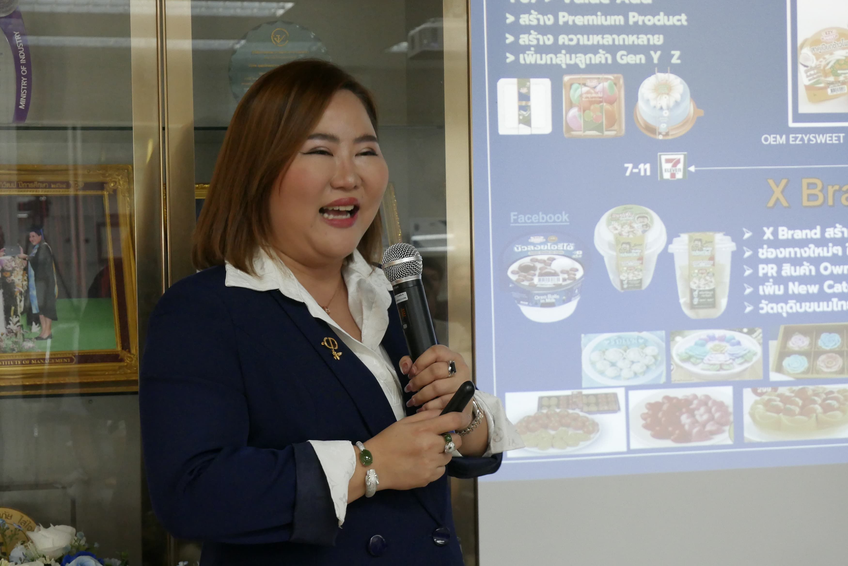 Woman in a blue blazer speaks into a microphone during a presentation with products displayed.