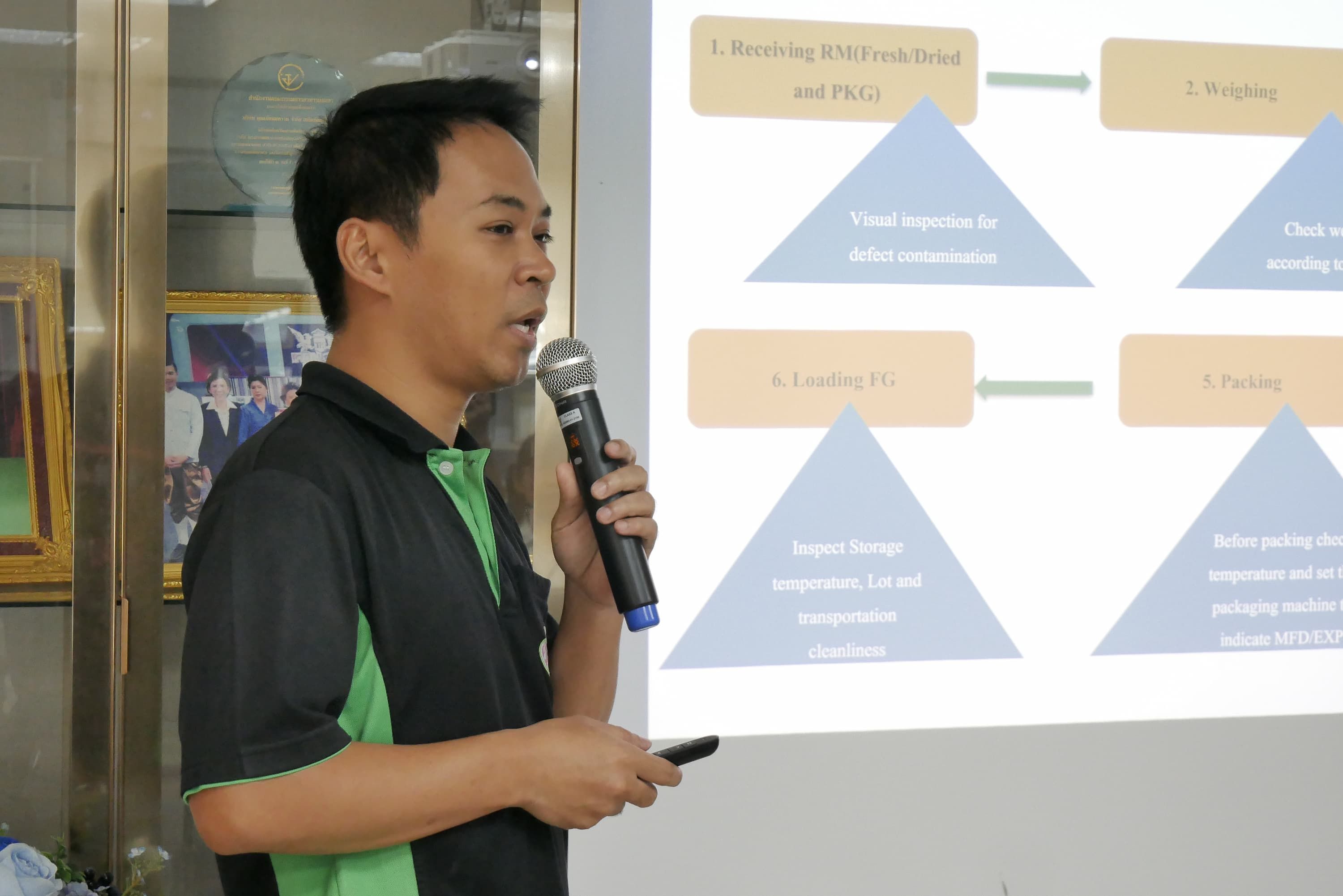 Man in green and black polo shirt speaks into microphone during a presentation with projected diagrams.