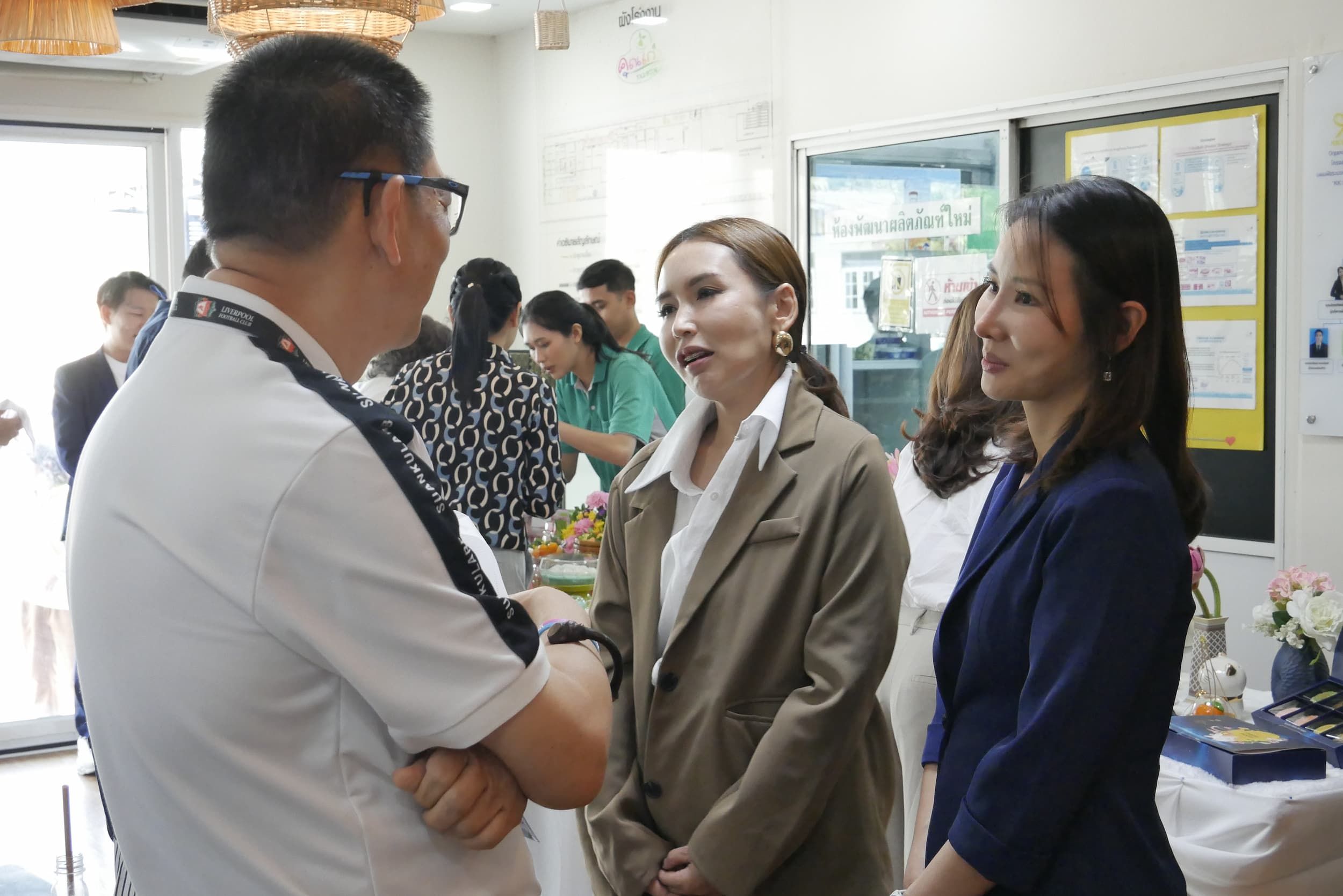 A man in glasses talks to two women in blazers in a professional indoor setting.