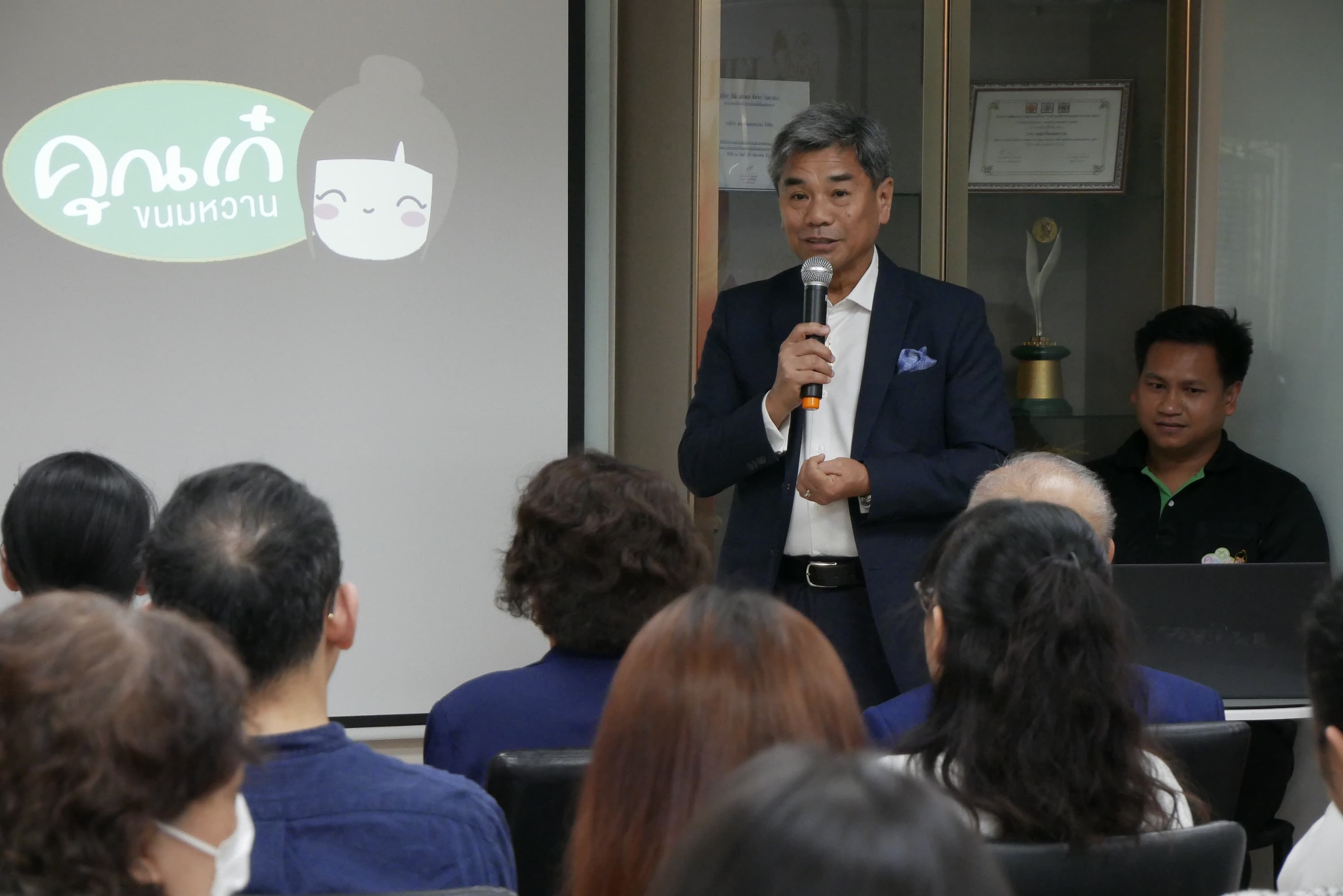 Man presents to an audience, a screen with a logo visible behind him.