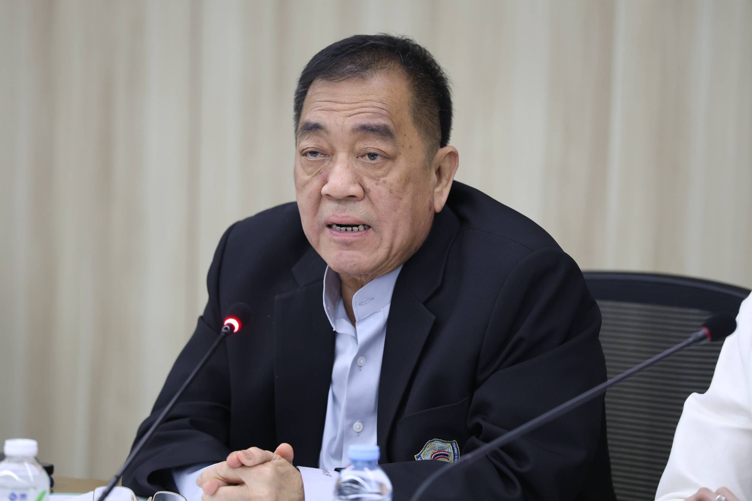 Older Asian man in dark suit speaks into a microphone at a formal table.