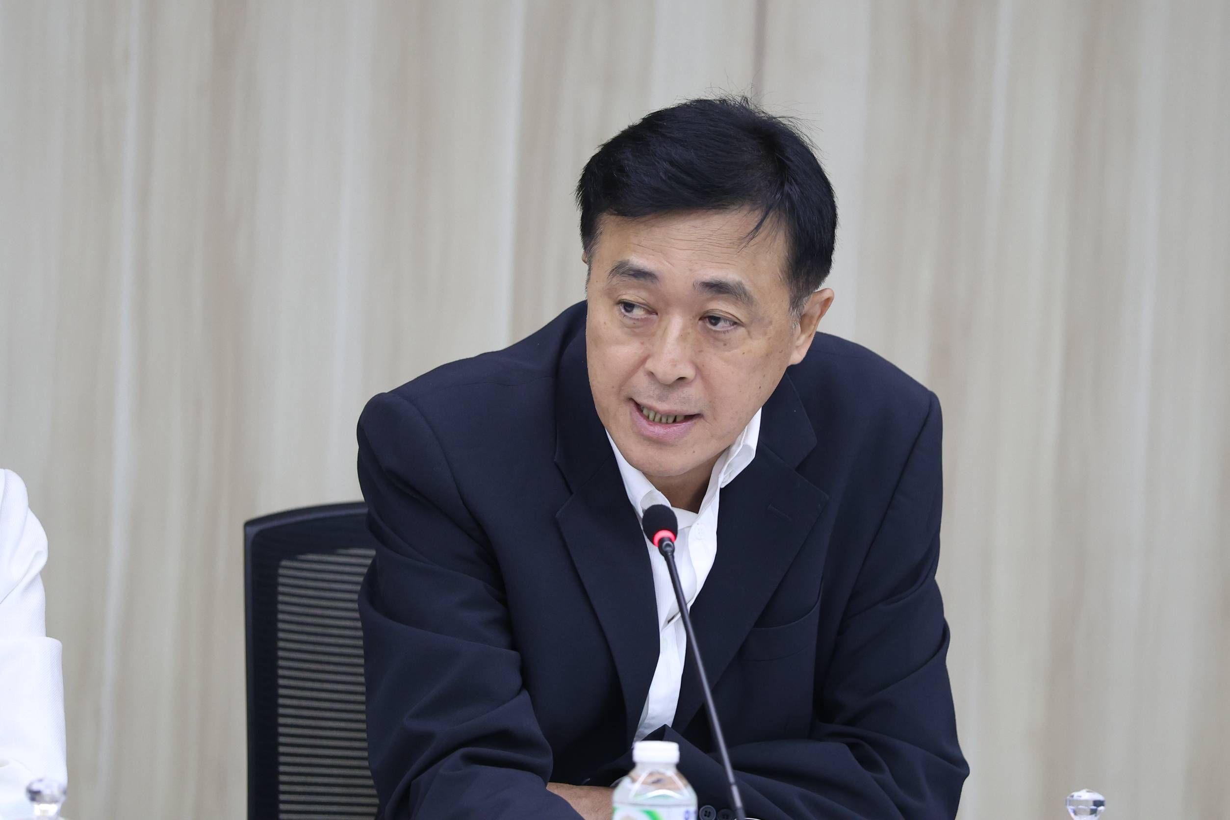 Man in dark suit speaking at a meeting with a microphone and water bottle.