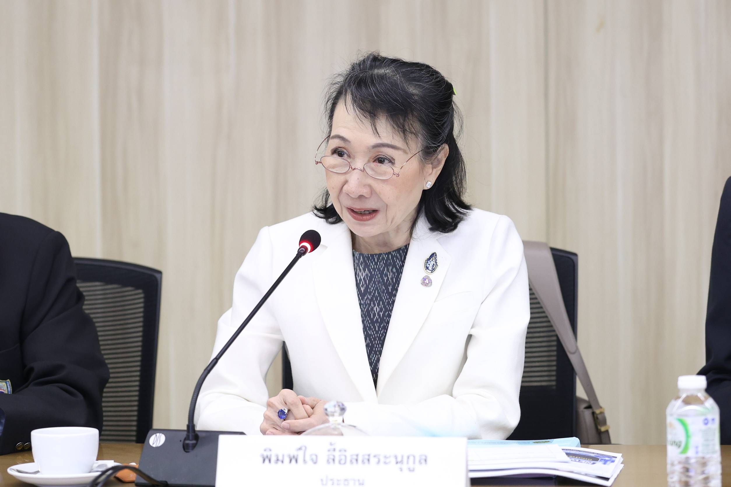 An Asian woman with glasses in a white blazer speaks into a microphone at a table.