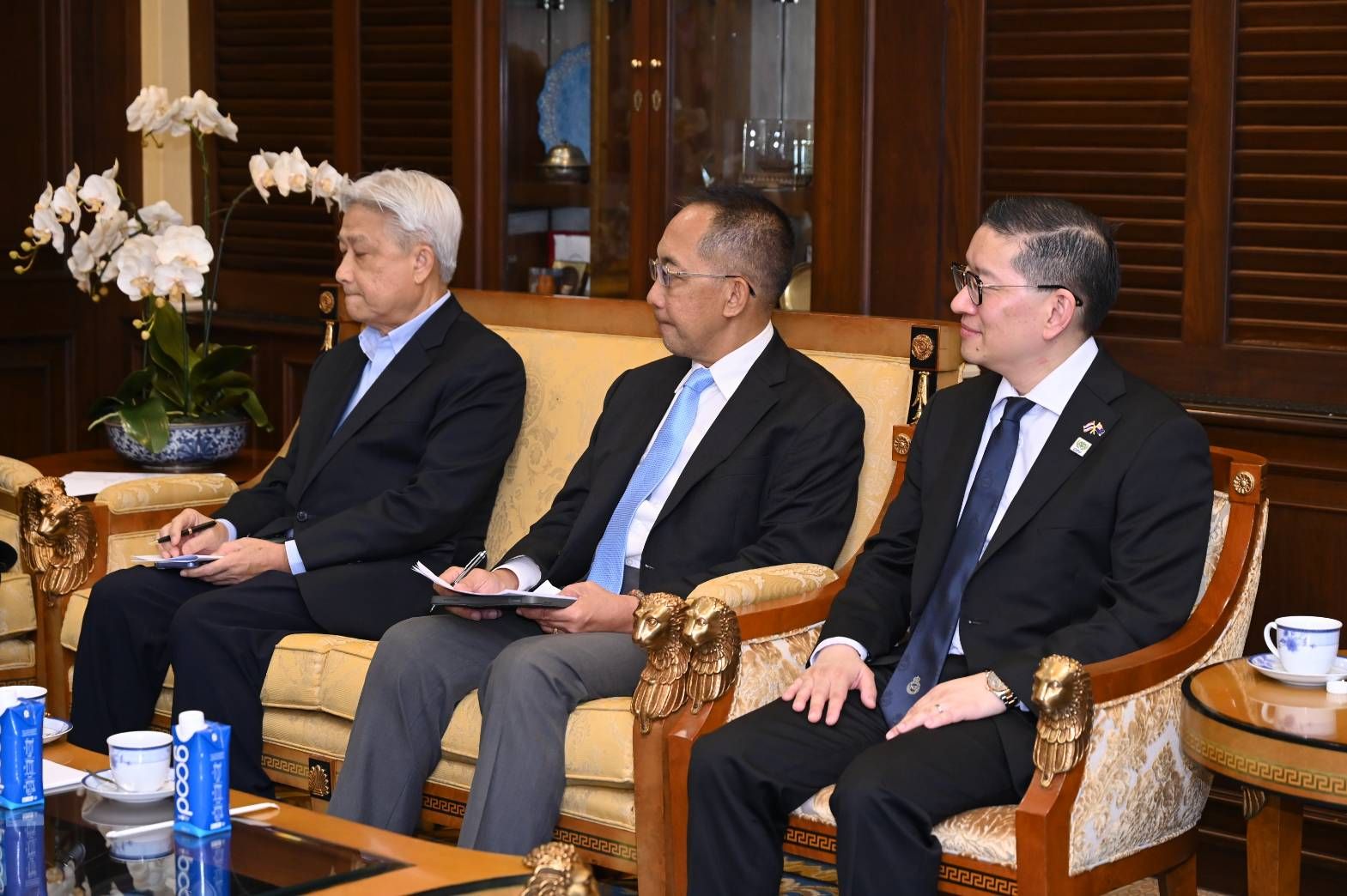 Three men in suits sit on a sofa in a formal room, attentively observing something.