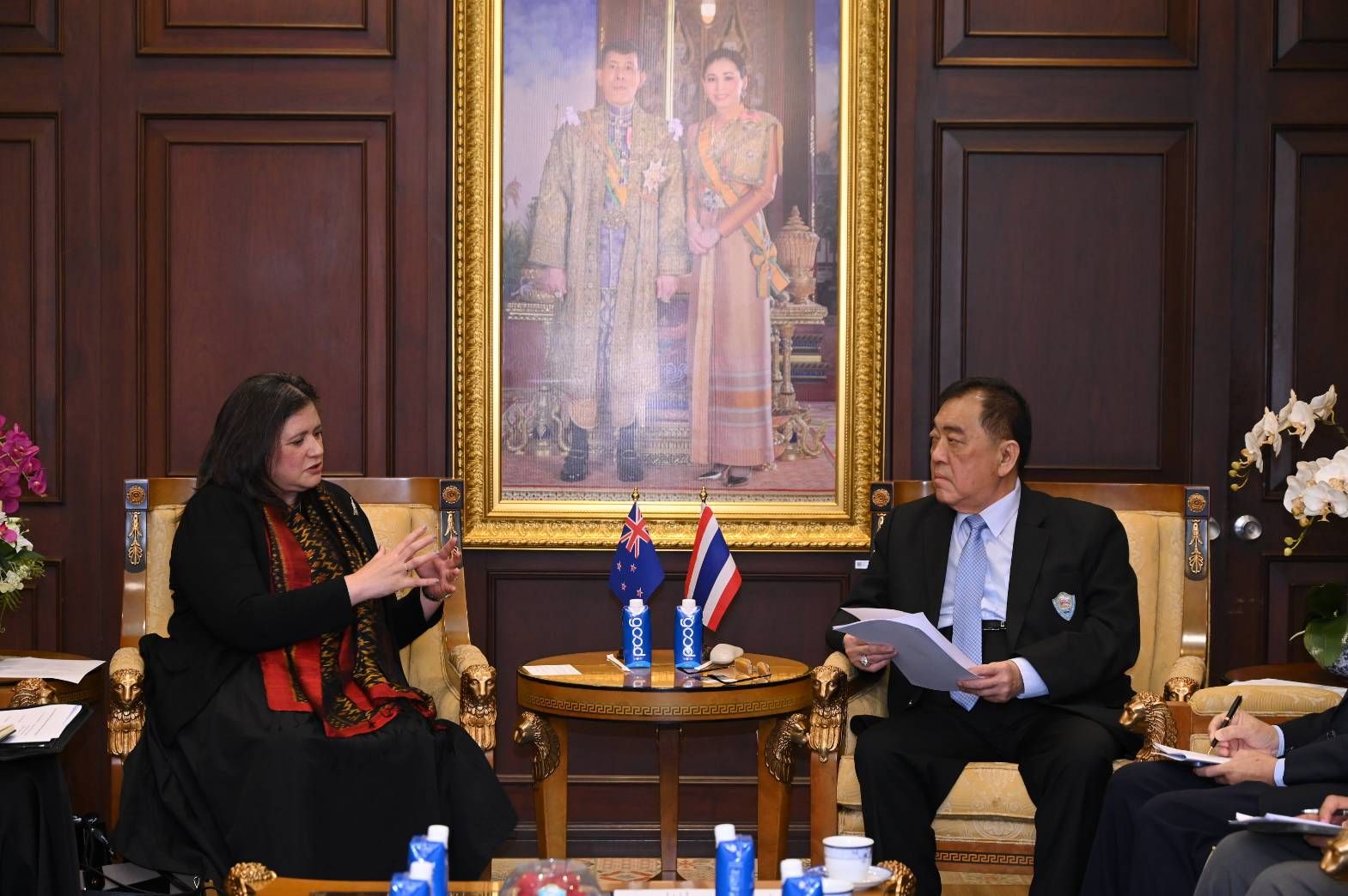 Two individuals discuss formally, a royal portrait hangs prominently in the background.