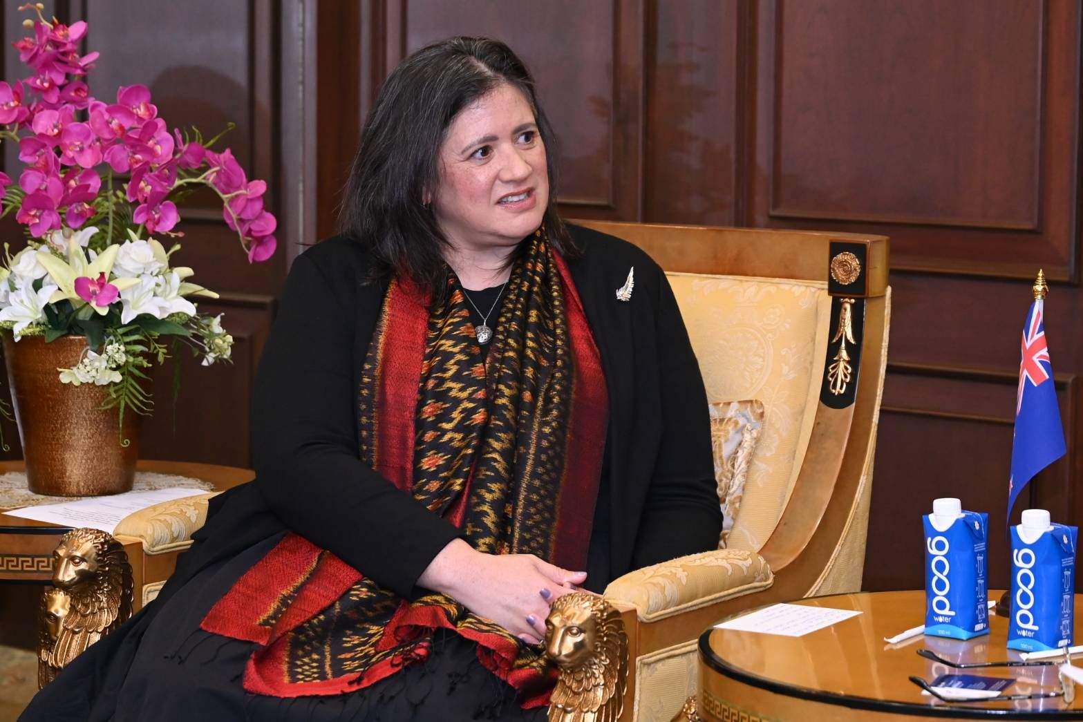 Woman in dark attire and patterned scarf seated formally, a flag and drinks nearby.
