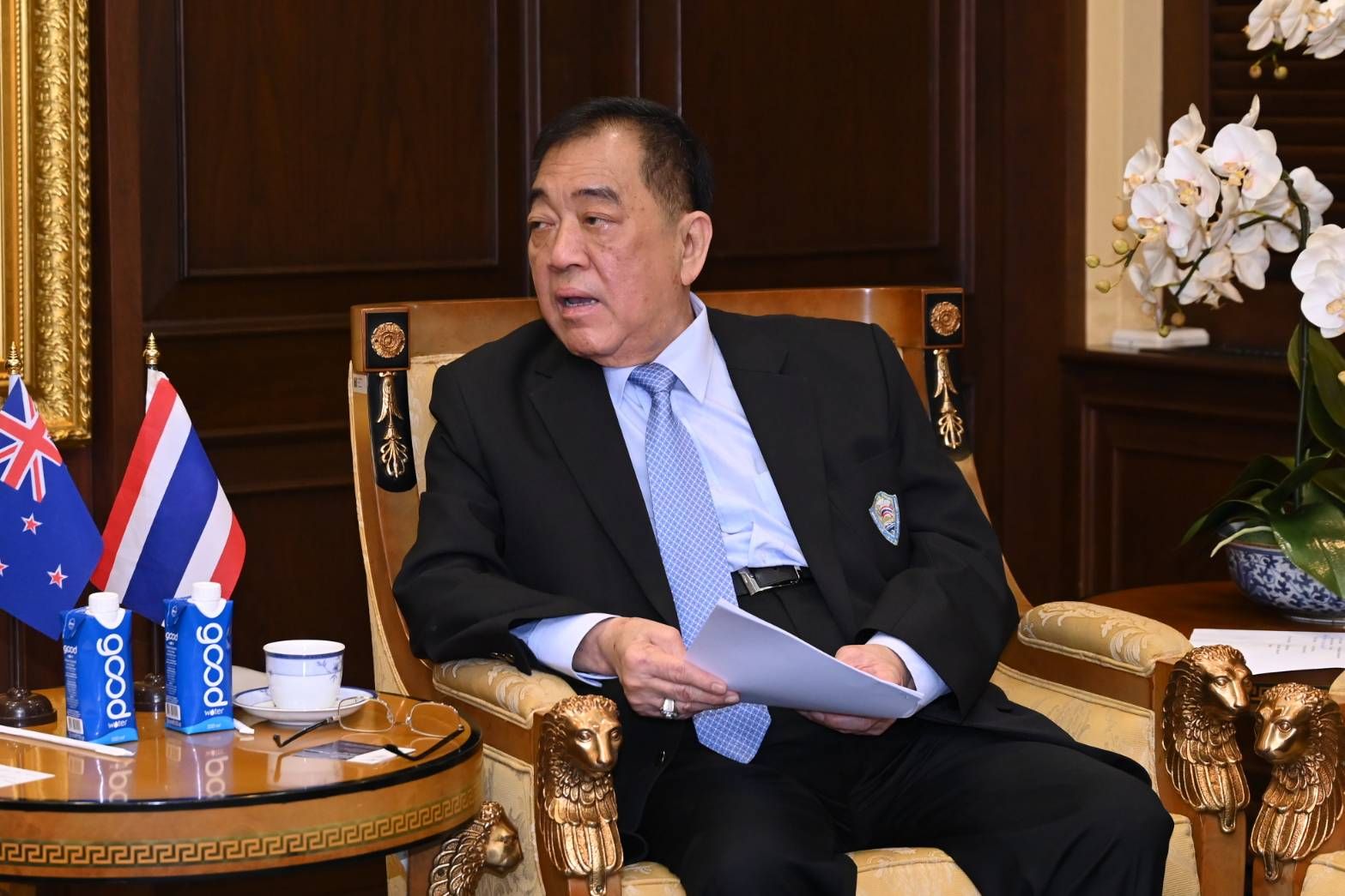 Older Asian man in a dark suit sits, holding papers, with flags in the background.