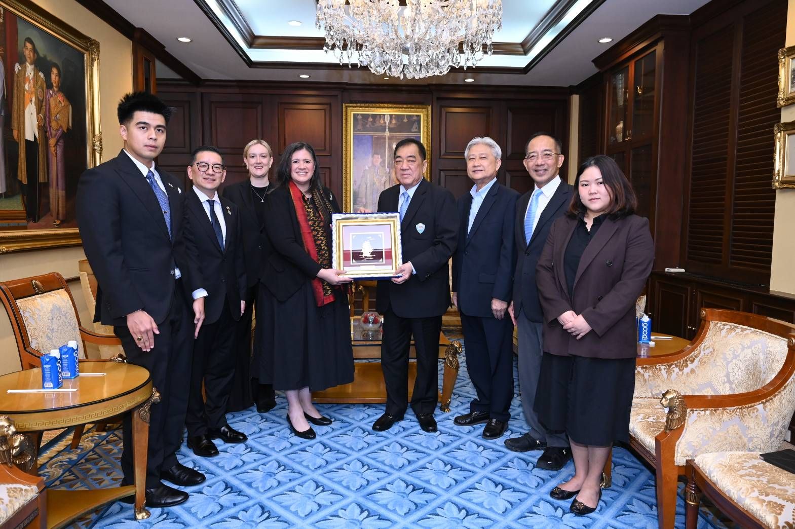 A group of delegates in a formal meeting room, a woman presents a framed gift to a man.