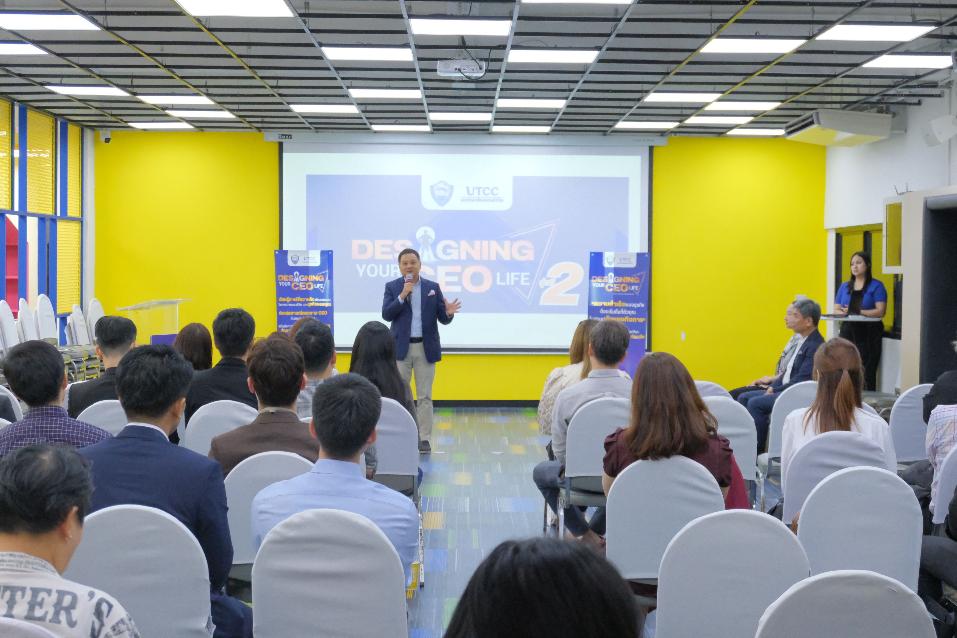 Man speaking at a conference to an attentive audience with a projector screen in the background.