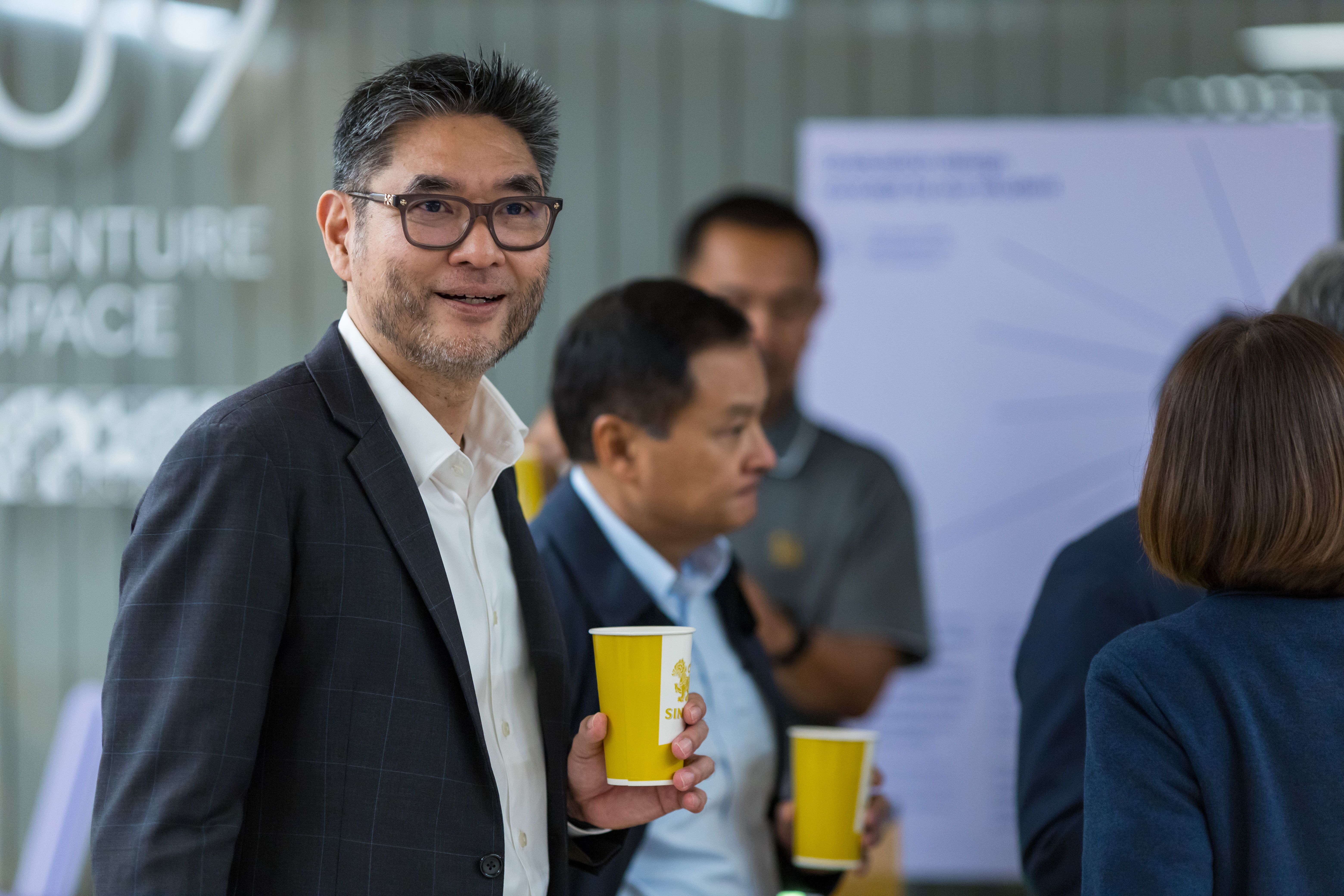 Smiling man in glasses and suit jacket holds yellow cup at an indoor event.