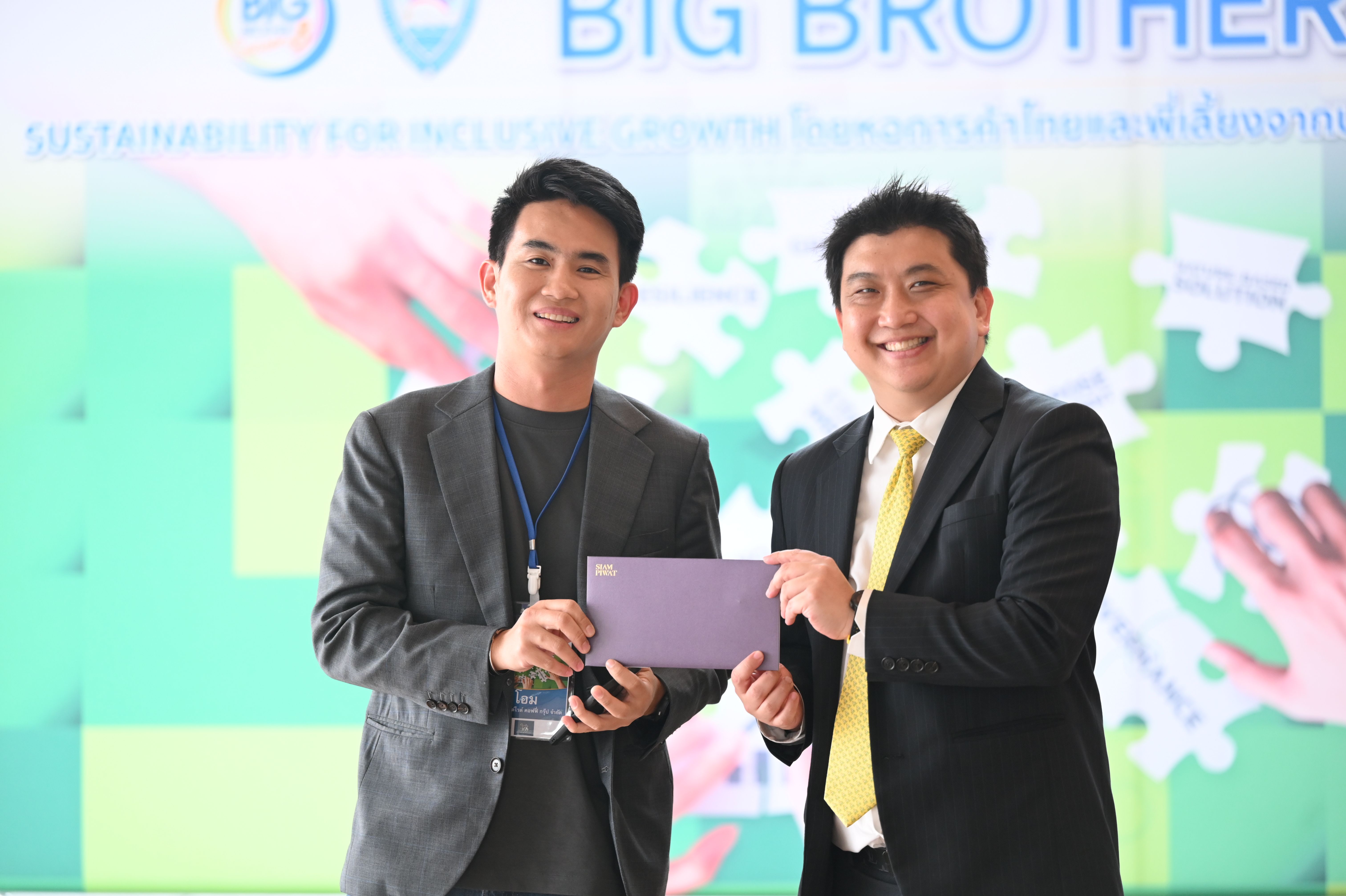 Two smiling men in suits jointly holding a purple envelope at a presentation.