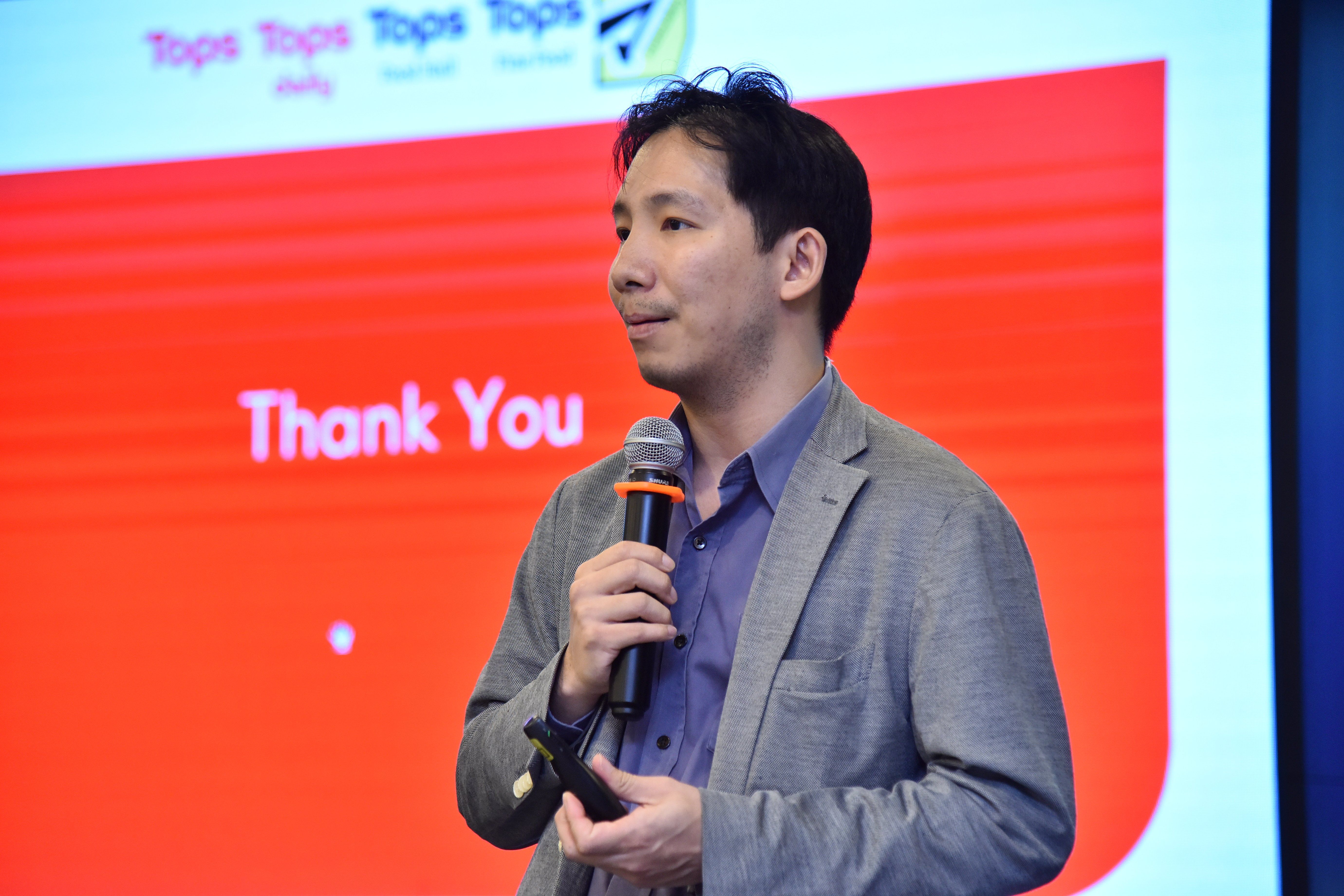 Man speaking into a microphone on stage with a red 'Thank You' background.