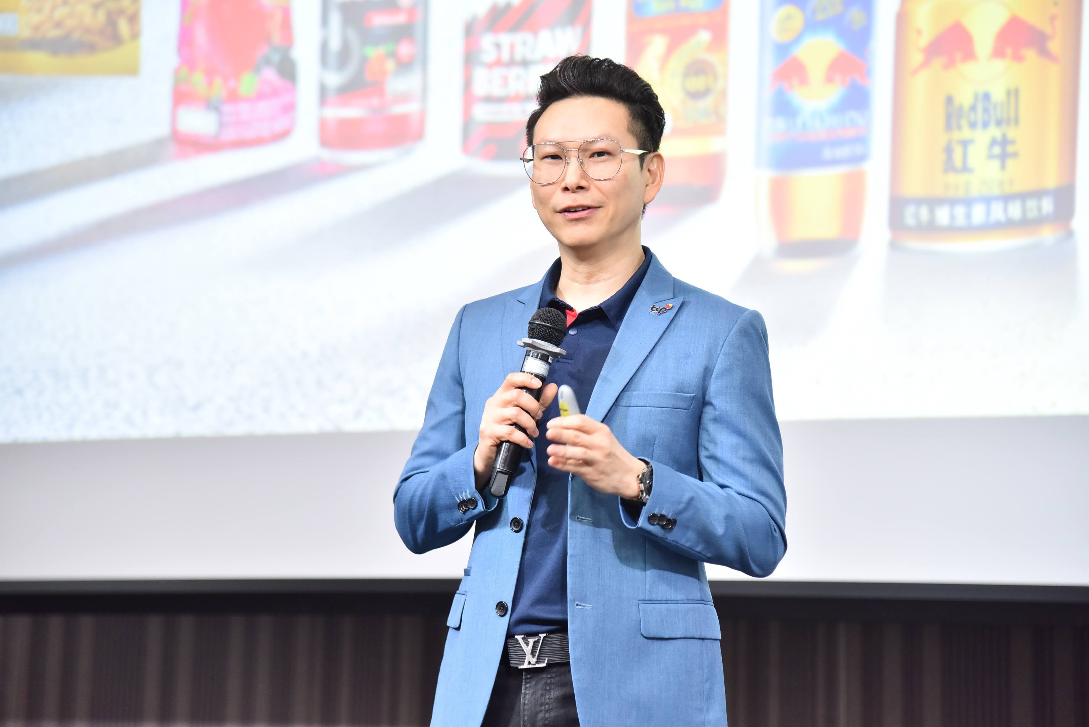 Man in blue suit and glasses speaks into a microphone with blurred energy drinks background.