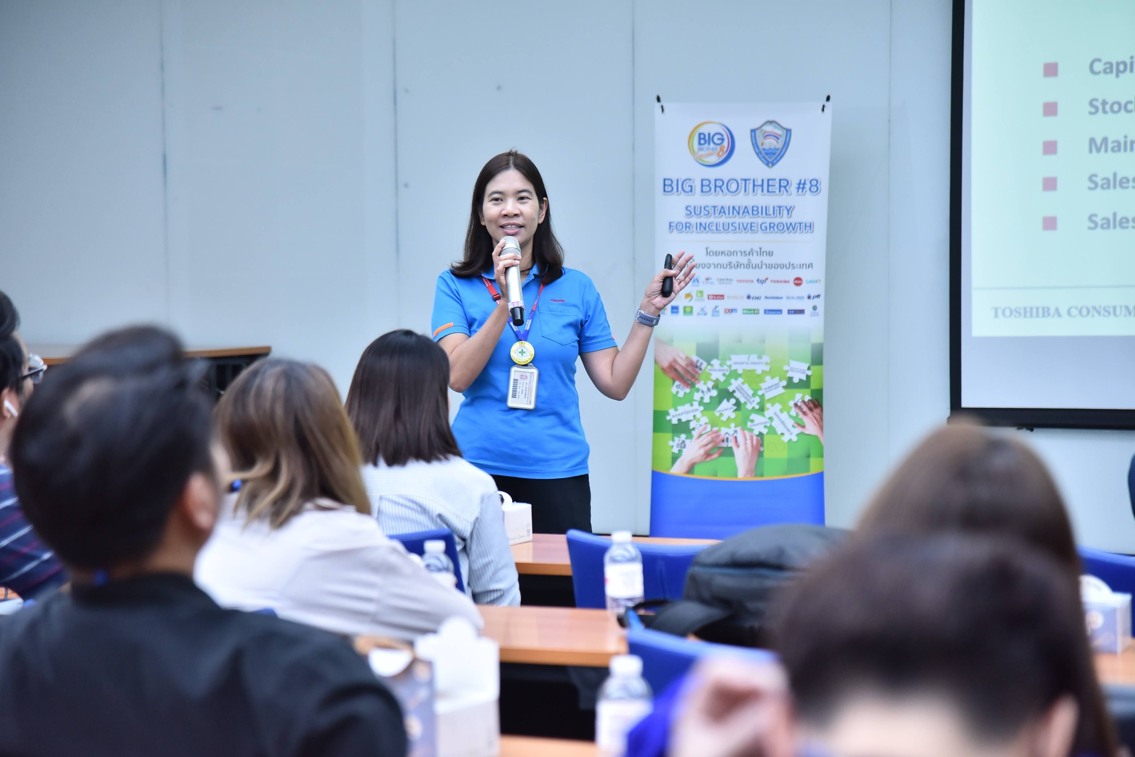 A woman in a blue shirt presents to an audience, gesturing in front of a screen.