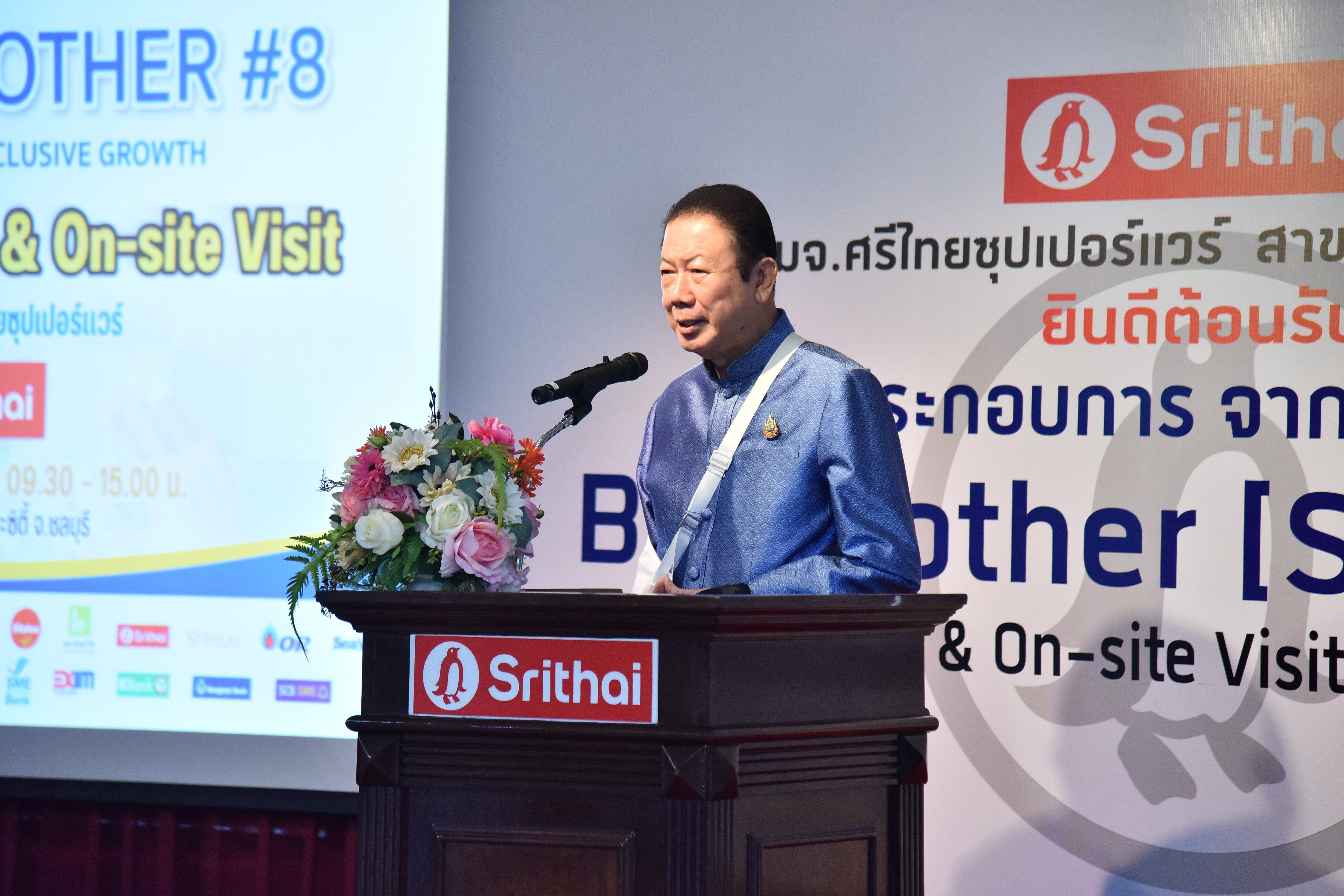 A man speaks at a podium with a microphone, a screen behind shows "Srithai" branding.