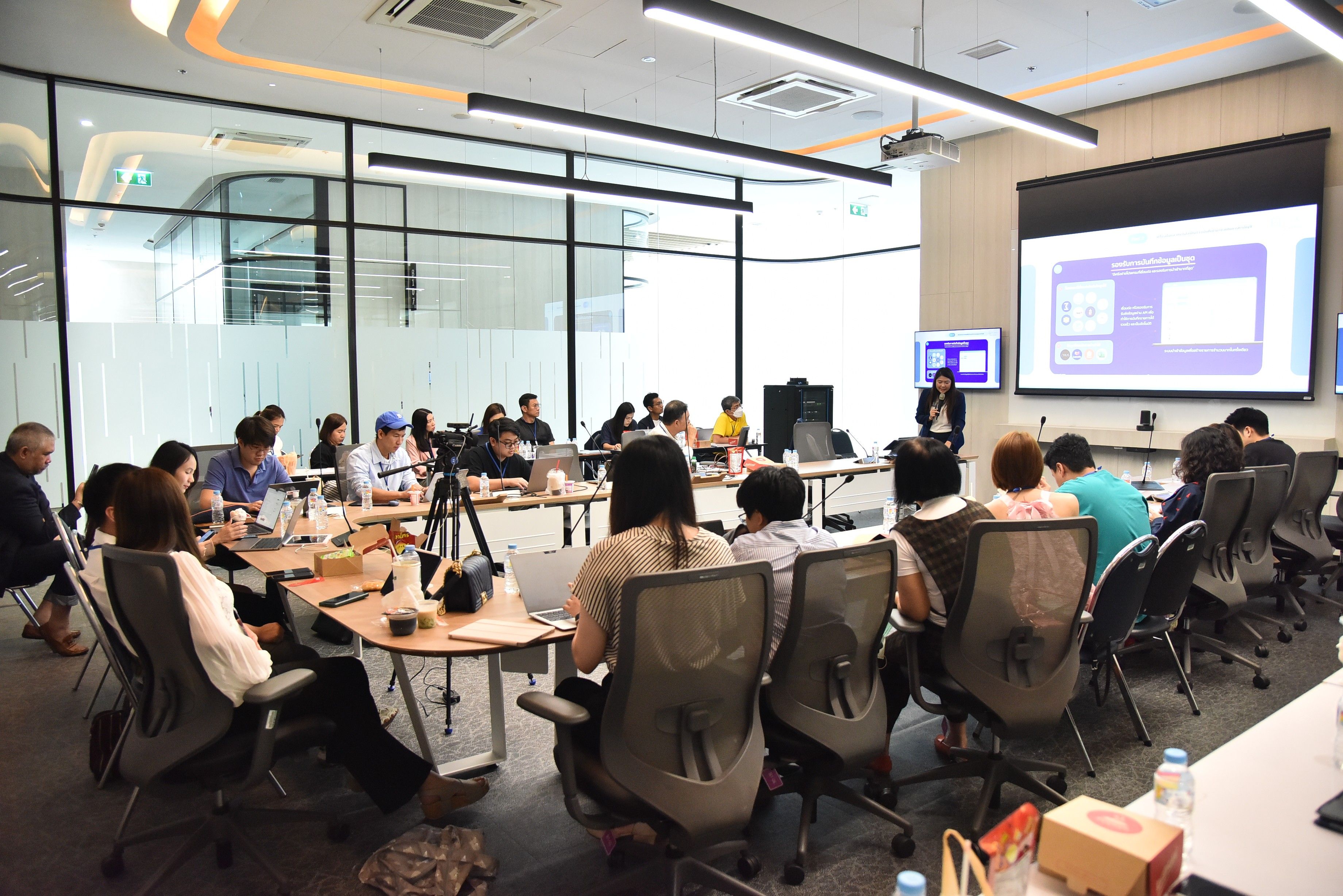 Attendees observe a presentation in a bright, modern conference room with glass walls.