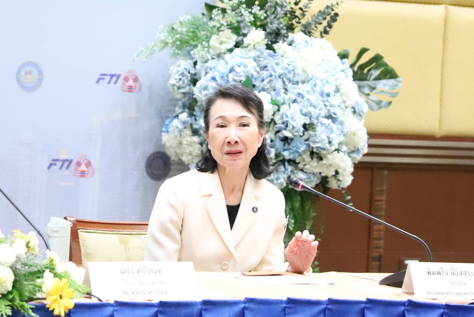 Professional woman in a cream suit speaks and gestures at a formal conference table.