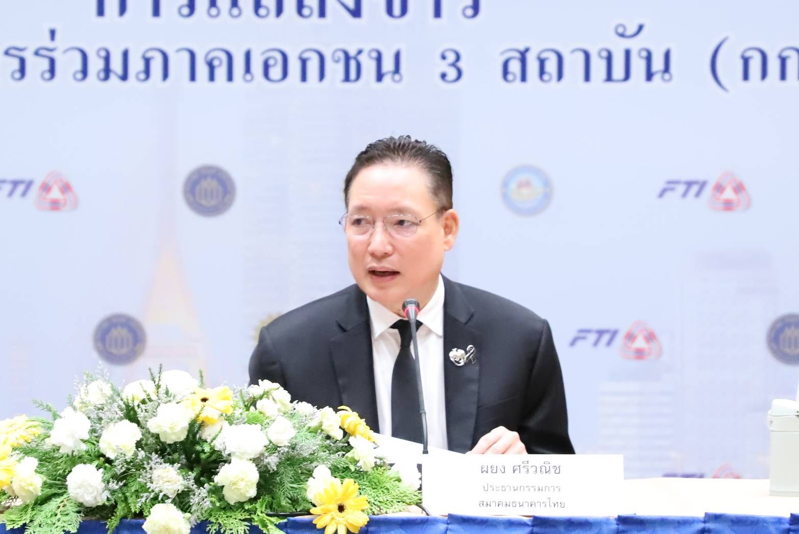Man in suit and glasses speaks at a podium with microphone and flowers.