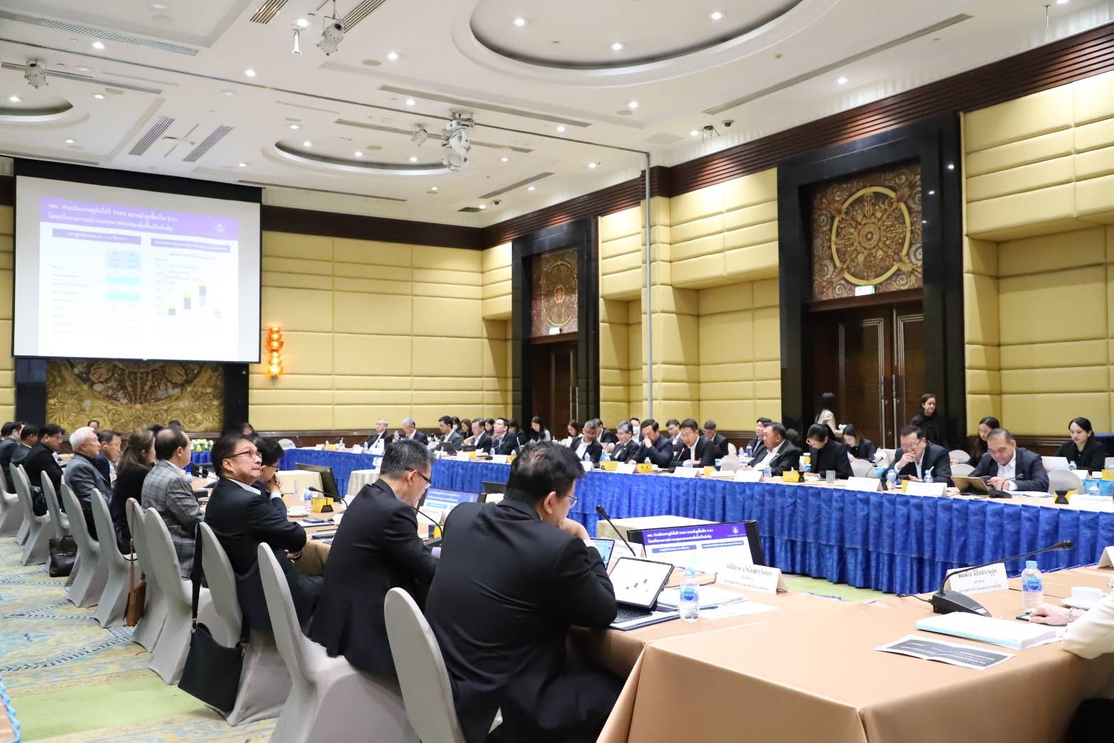 Business conference attendees watch a presentation on a large screen in a meeting room.